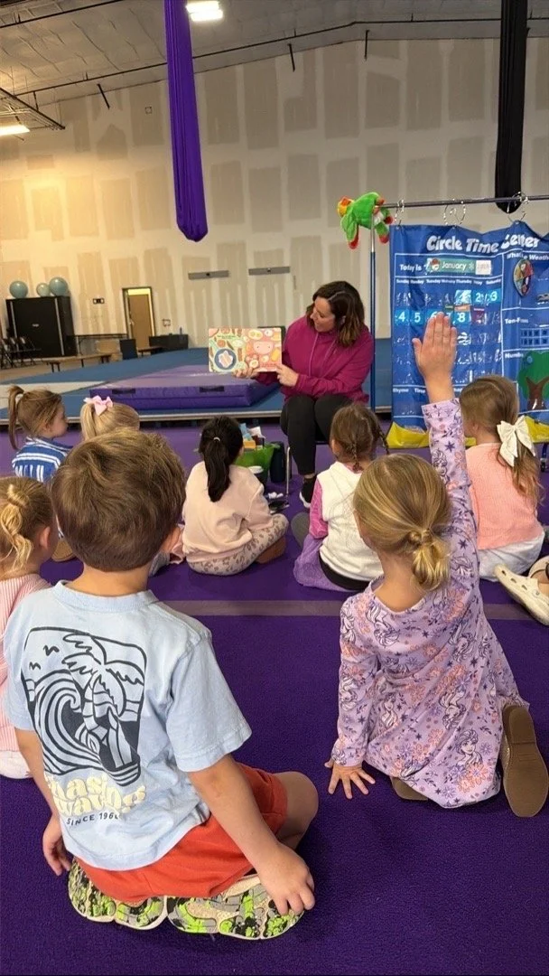A woman reading a book to a group of children seated on a purple mat in a classroom or activity room, with one child raising her hand.
