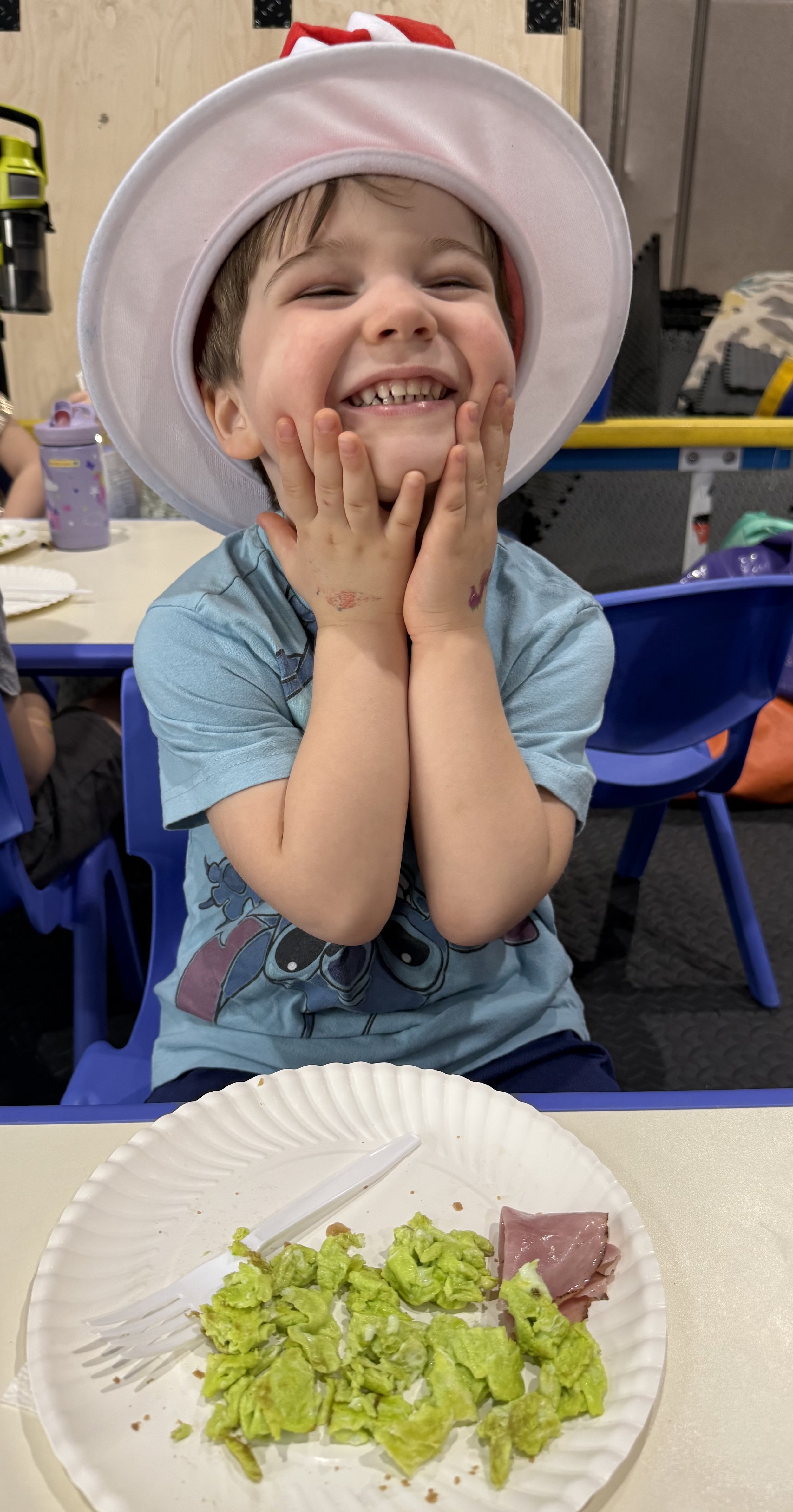 A young boy smiling happily with his hands on his cheeks, wearing a white holiday hat with red accents, sitting at a table with a paper plate in front of him that has some lettuce, a slice of ham, and a plastic fork.