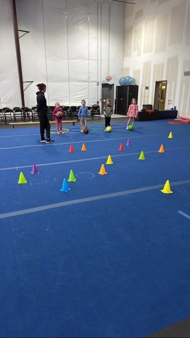 Four young children standing on a blue gym floor, each with a green ball, in a gym or sports facility with cones arranged in a pattern, and a woman instructing them.
