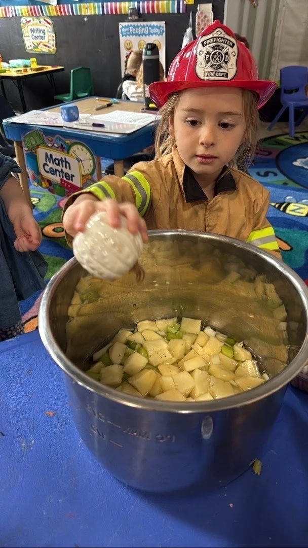 A young girl dressed as a firefighter with a red firefighter helmet, in a classroom, pouring chopped vegetables into a large metal pot during a cooking activity.