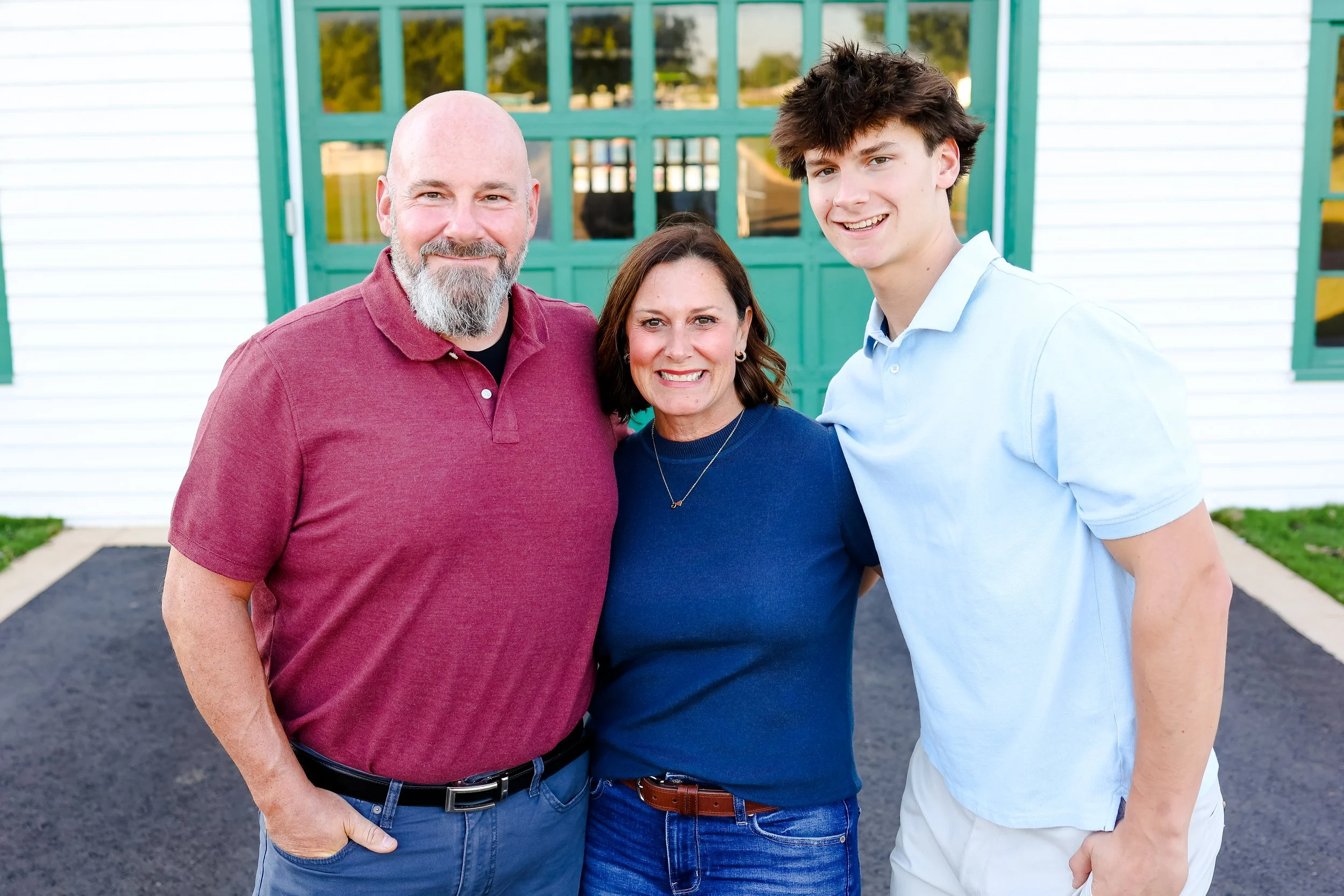 A smiling family of three standing outside in front of a green garage door, with a white house behind them. The family includes a man with a beard in a red polo shirt, a woman in a blue shirt, and a teenage boy in a light blue shirt.