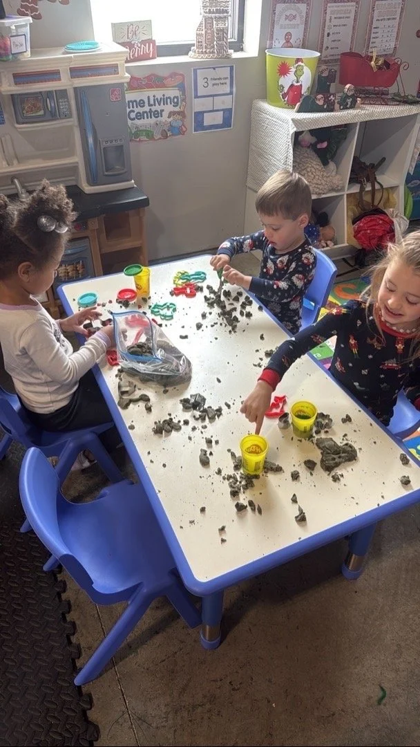 Three children playing with modeling clay and cookie cutters at a white table in a classroom.