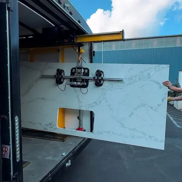 A large slab of white marble with gray veining is being moved or installed from a truck. The marble is secured by a robotic arm or lifting device, and a person is holding the slab on the right side.