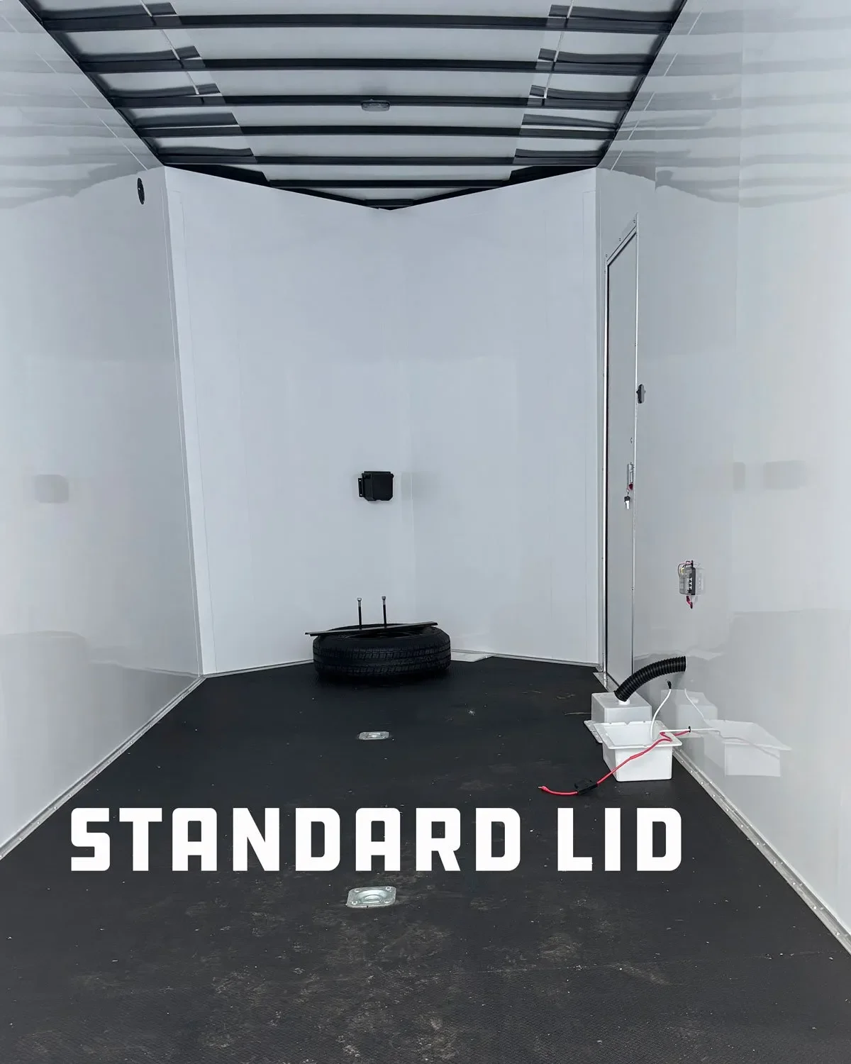 Empty interior space with a black rubber floor, white walls, a door with a lock, a tire with tools on top, and a white electrical box with wiring on the right side. Text overlays the image reading 'STANDARD LID'.