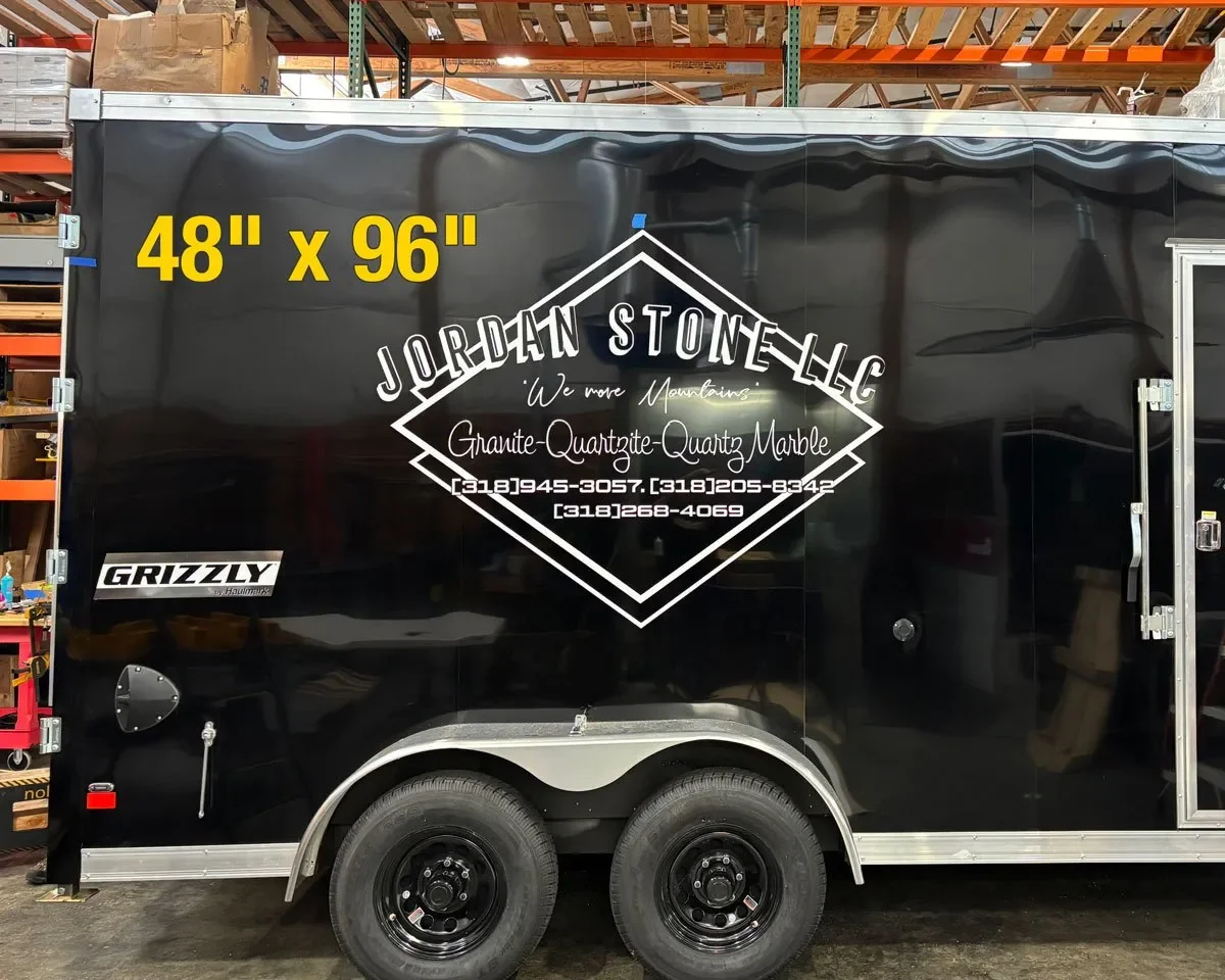 Black trailer with white and yellow text advertising Jordan Stonellc granite and marble services, with a phone number, and a logo for Grizzly. The trailer is in a warehouse or workshop.