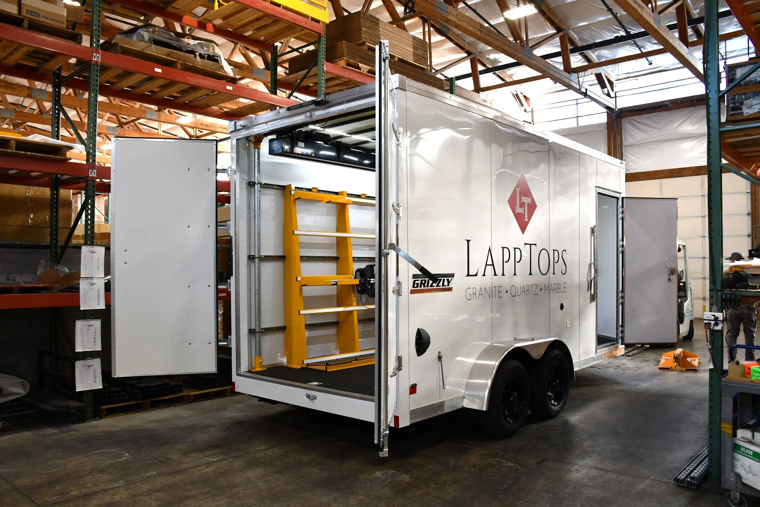A mobile workshop trailer labeled 'LAPTOPS' with the words 'GRANITE, QUARTZ, MARBLE' and a logo with a diamond shape, parked inside a warehouse with metal shelving and wooden pallets overhead.