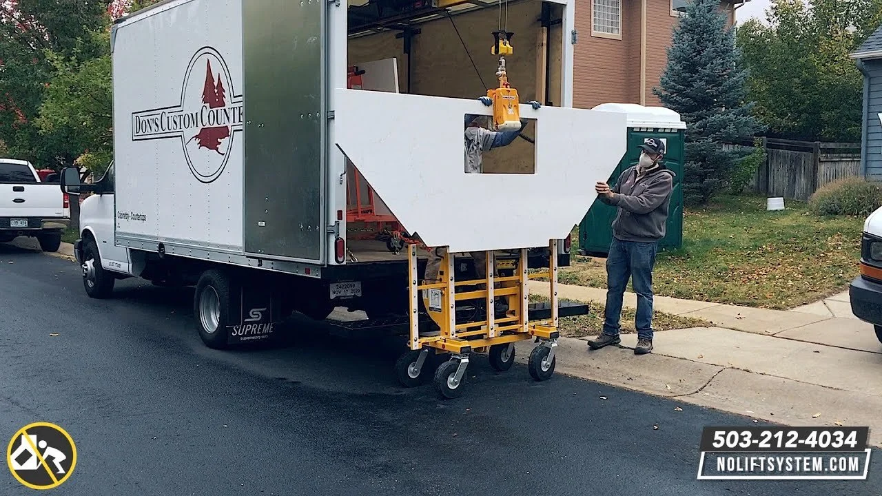 A worker in a gray hoodie and face mask standing outside next to a white truck with a sign that reads 'Don's Custom Counter'. The truck has a large white cabinet with a cutout window being installed, with a yellow lifting device attached to it. The scene is in a residential neighborhood with houses, trees, and parked cars.
