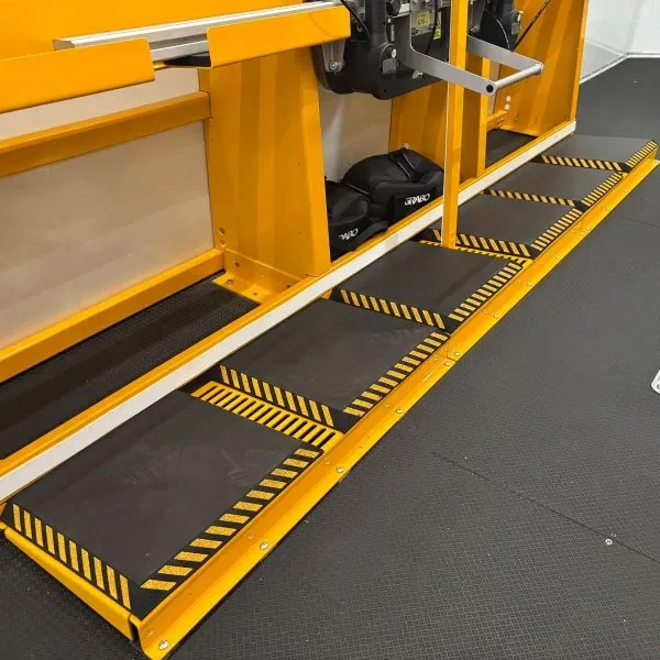 A yellow safety transfer platform with five non-slip black mats, designed for moving wheelchairs or gurneys, located next to a vertical yellow metal structure with black and yellow striped caution markings on the floor.
