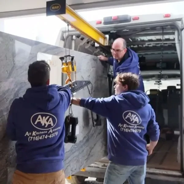 Three workers in blue hoodies installing a large stone slab using a crane on a truck. One worker is controlling the crane while two others assist.
