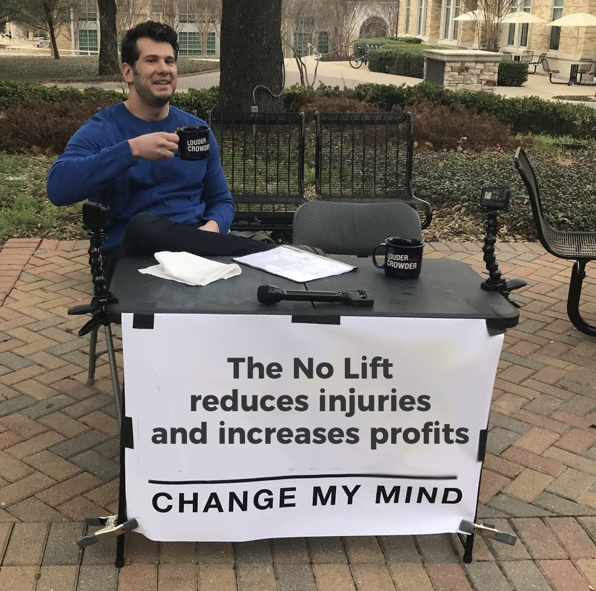 Man sitting at a table with a sign that reads, "The No Lift reduces injuries and increases profits. Change My Mind." He is holding a mug that says, "Louder Crowder," and there are microphones on the table, with chairs and benches in the background.