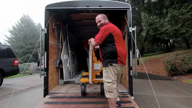 A man in a red and black shirt and khaki shorts standing inside the open back of a black moving trailer, smiling and holding a hand truck with books.