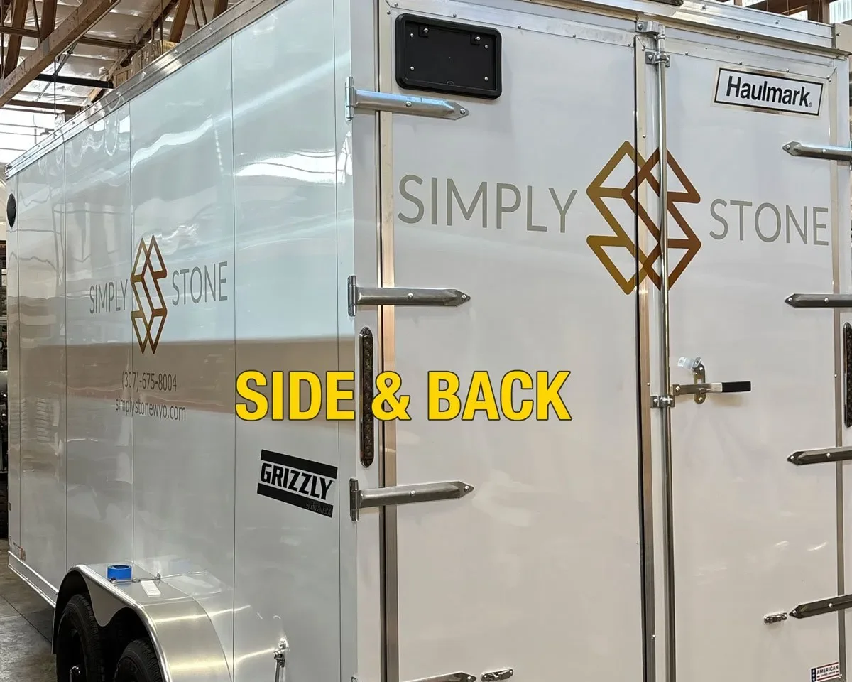 A white trailer with gold and black text and logos, parked inside a building. The trailer has a door with metal handles and latches, and the side and back are shown. The text on the trailer reads "SIMPLY STONE" and