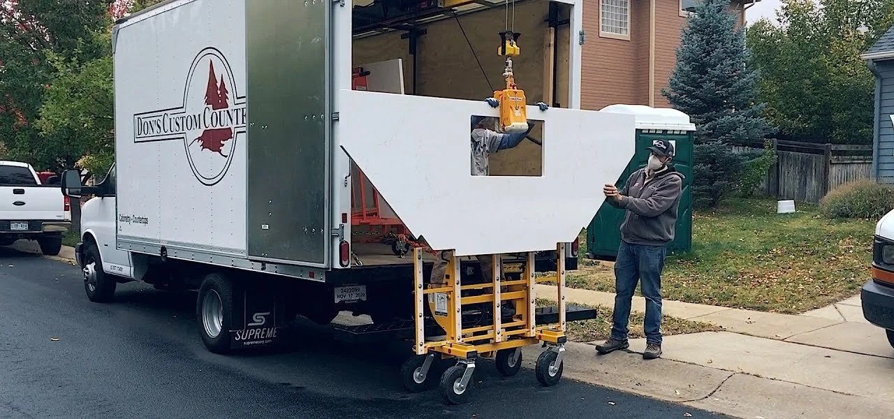 A person wearing a mask is standing on the sidewalk next to a mobile workshop truck with the logo 'Don's Custom Counters.' The truck has a partially painted white counter or workspace extension. The scene is in a residential neighborhood with houses, trees, and parked cars.