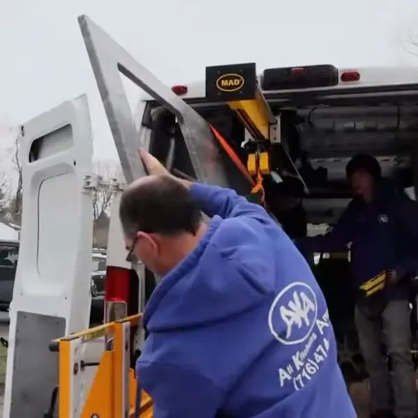 Two workers loading equipment onto a truck from a utility van on a cloudy day. One worker, wearing glasses and a blue hoodie, is handling a yellow piece of equipment, while another worker in the background assists.