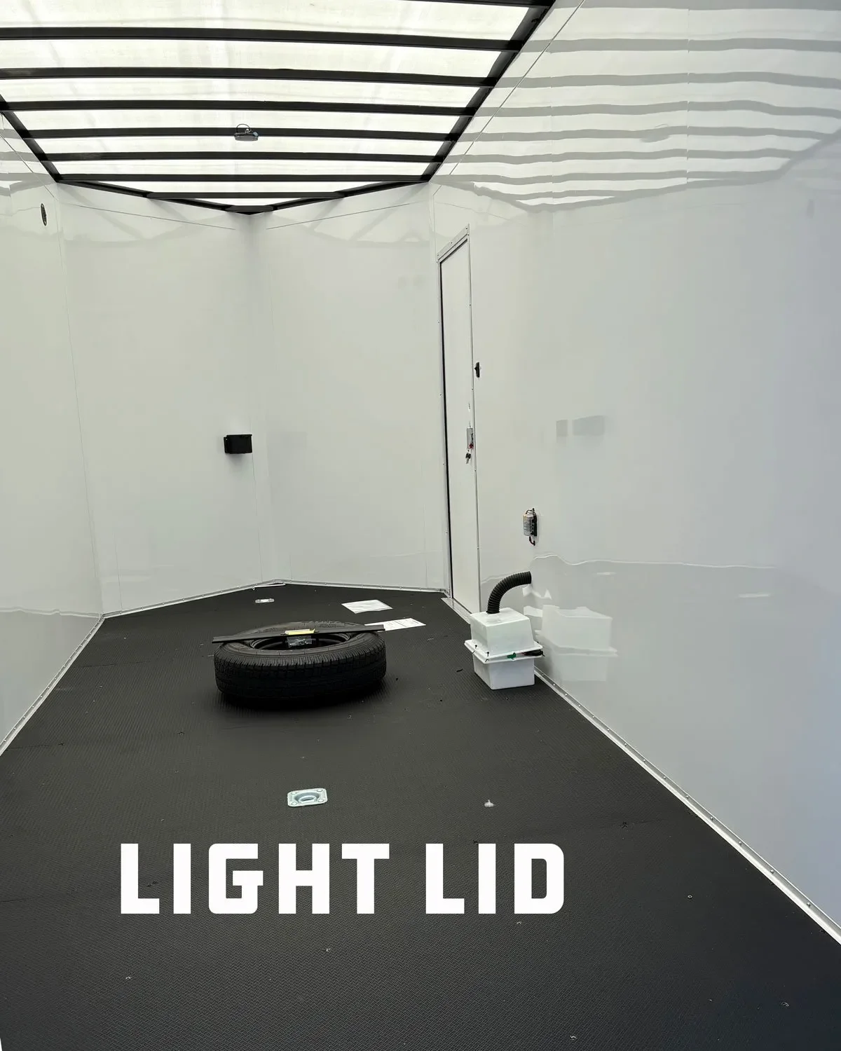Empty, clean, lighted interior of a small room or trailer with a black floor, white walls, and a translucent ceiling with black framing. Contains a tire, a vent or outlet, some papers, and a small equipment setup with a filter and hose.