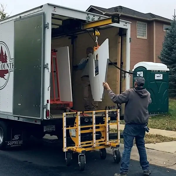 Two men are unloading large white panels from a moving truck parked on a residential street. One man inside the truck is holding a panel, while the other outside is assisting, standing on the ground. There is a green portable toilet and a house with brown siding in the background.