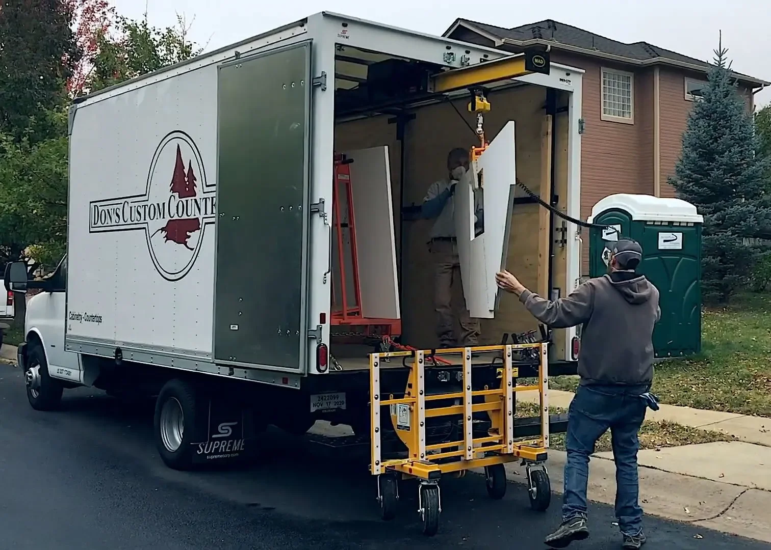 Workers loading or unloading white cabinets from a moving truck on a residential street; the truck has a logo that says 'Don's Custom Counter' and is parked near a house with a brown exterior and a small blue spruce tree.