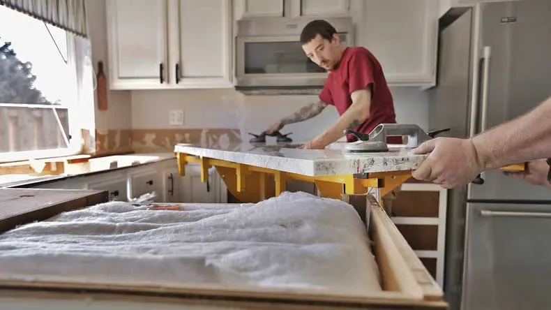 Two men are working together on a kitchen renovation, installing a new countertop, with one using a power drill and the other handling a power sander.