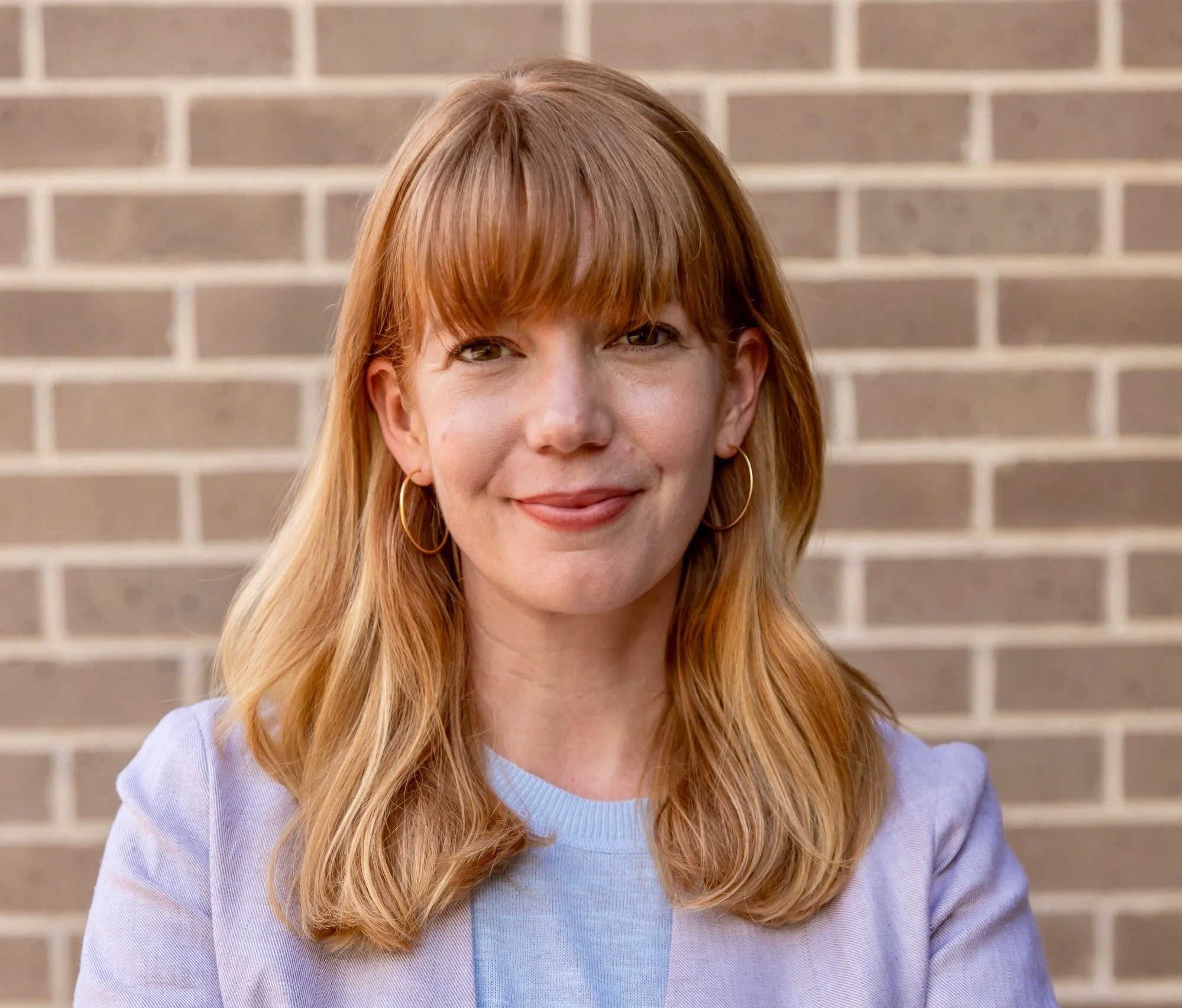 A woman with red hair and bangs, wearing hoop earrings and a light blue shirt, standing in front of a brick wall.