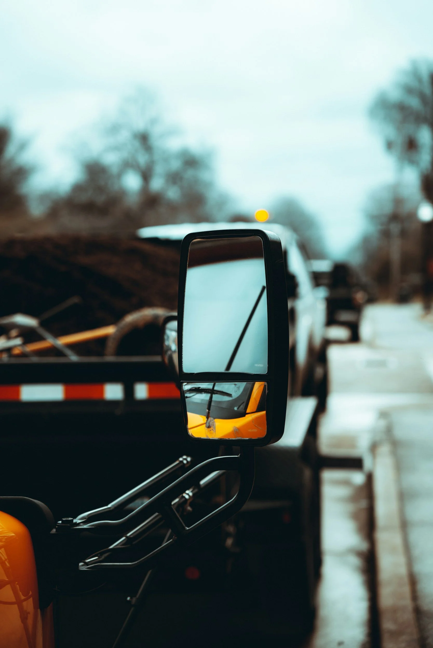 Close-up of a motorcycle's side mirror reflecting a yellow object, with a blurred background of trees and a vehicle, possibly a truck or van, on a cloudy day.