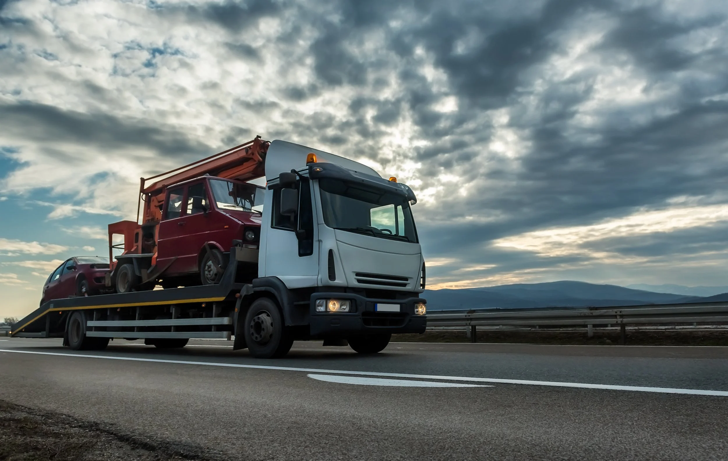 A flatbed truck transporting a red car and a red vintage vehicle on a highway with a cloudy sky and mountains in the background.