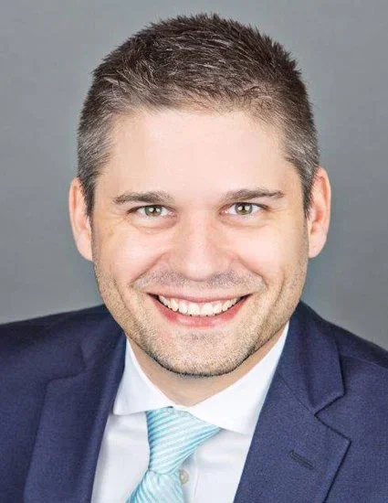 Close-up portrait of a smiling man in a suit and tie, with short brown hair and light skin, against a plain gray background.