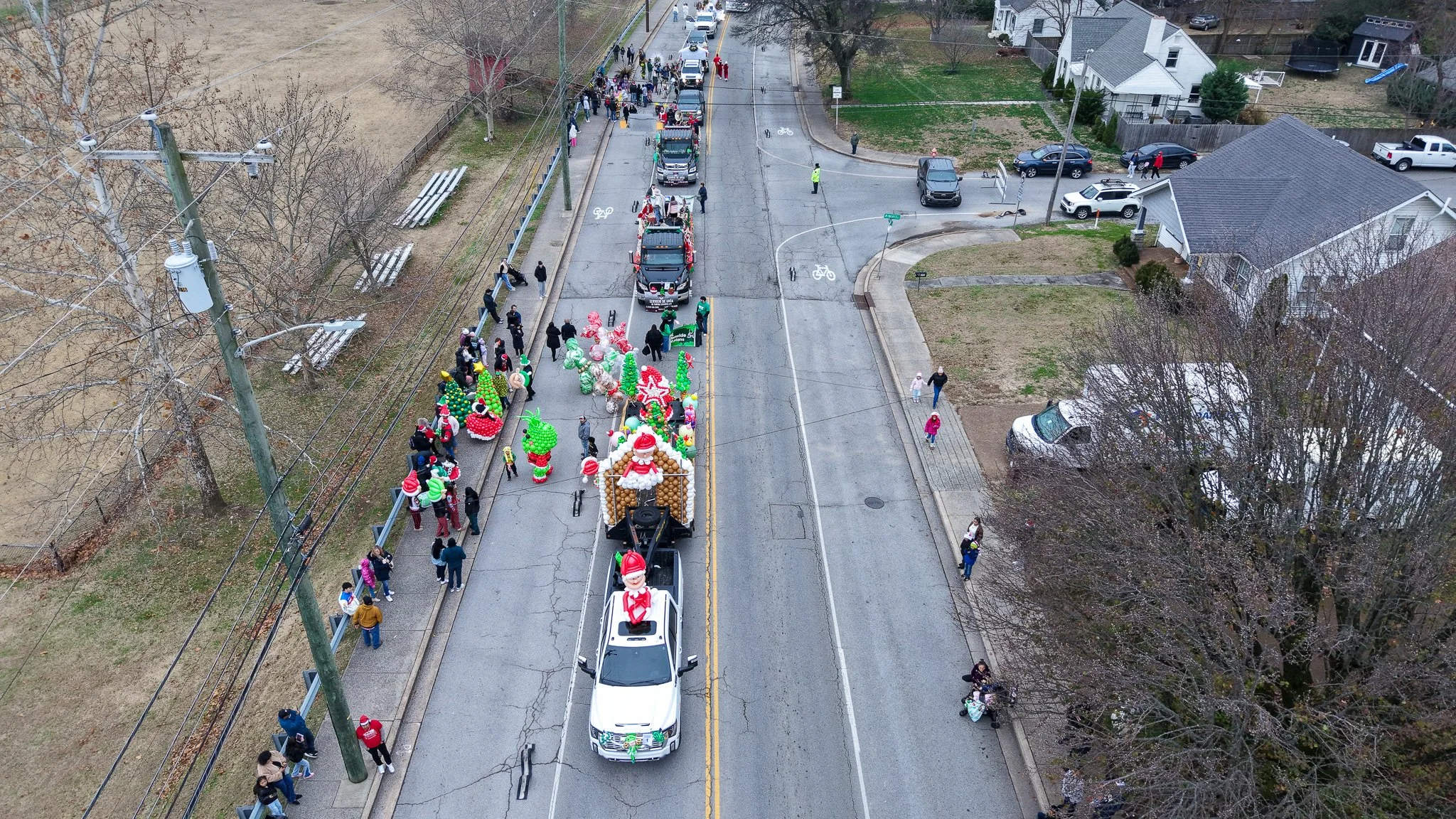 A parade with trucks decorated with Christmas-themed balloons and ornaments, moving down a street lined with spectators, some taking photos and watching the parade, with leafless trees and houses visible on the sides.