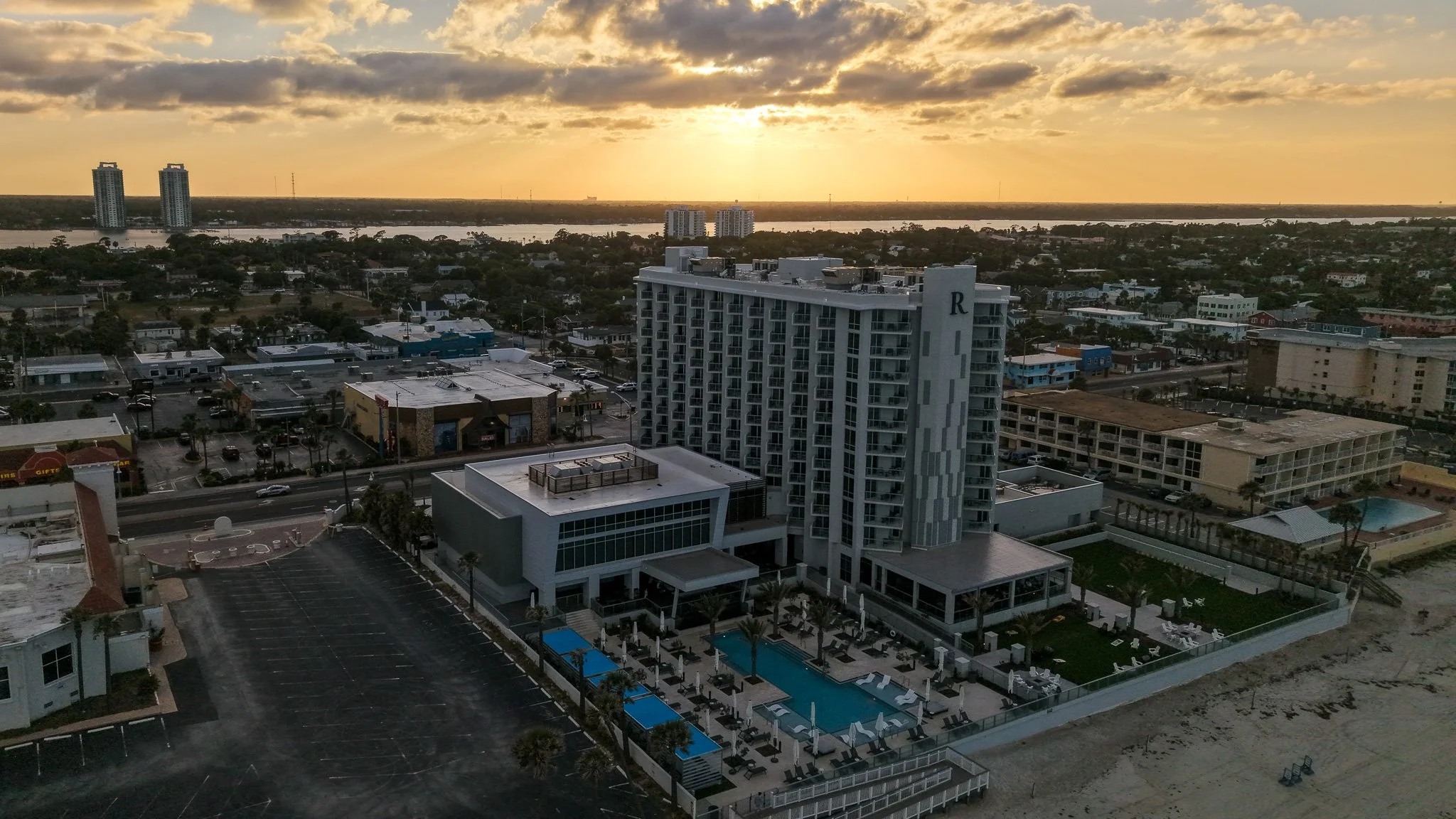 Aerial view of a beachfront hotel with swimming pool, surrounded by buildings and parking lot, during sunset.