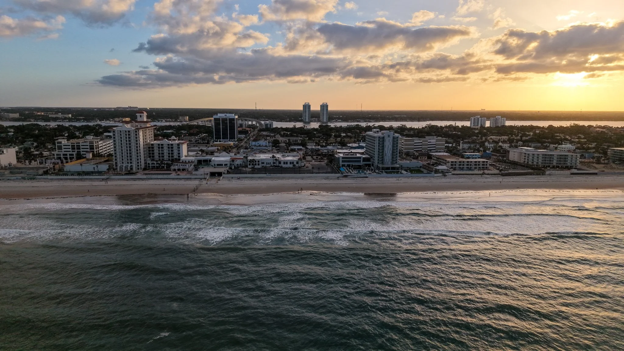 Aerial view of a beach at sunset with waves crashing on the shore, and a city skyline with mid-rise buildings and two tall towers in the background.