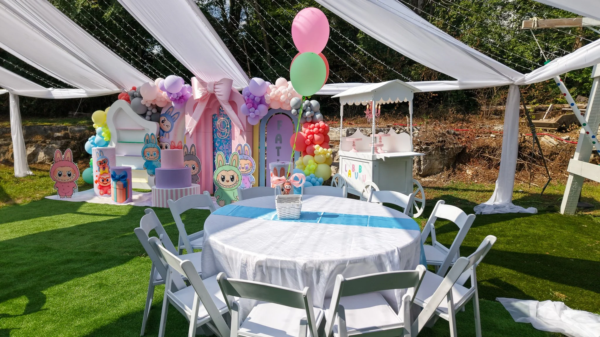 Decorated outdoor birthday party setup with pastel-colored balloons, cartoon character cutouts, a cake on a table, and a food stand under a white canopy on a grassy area.