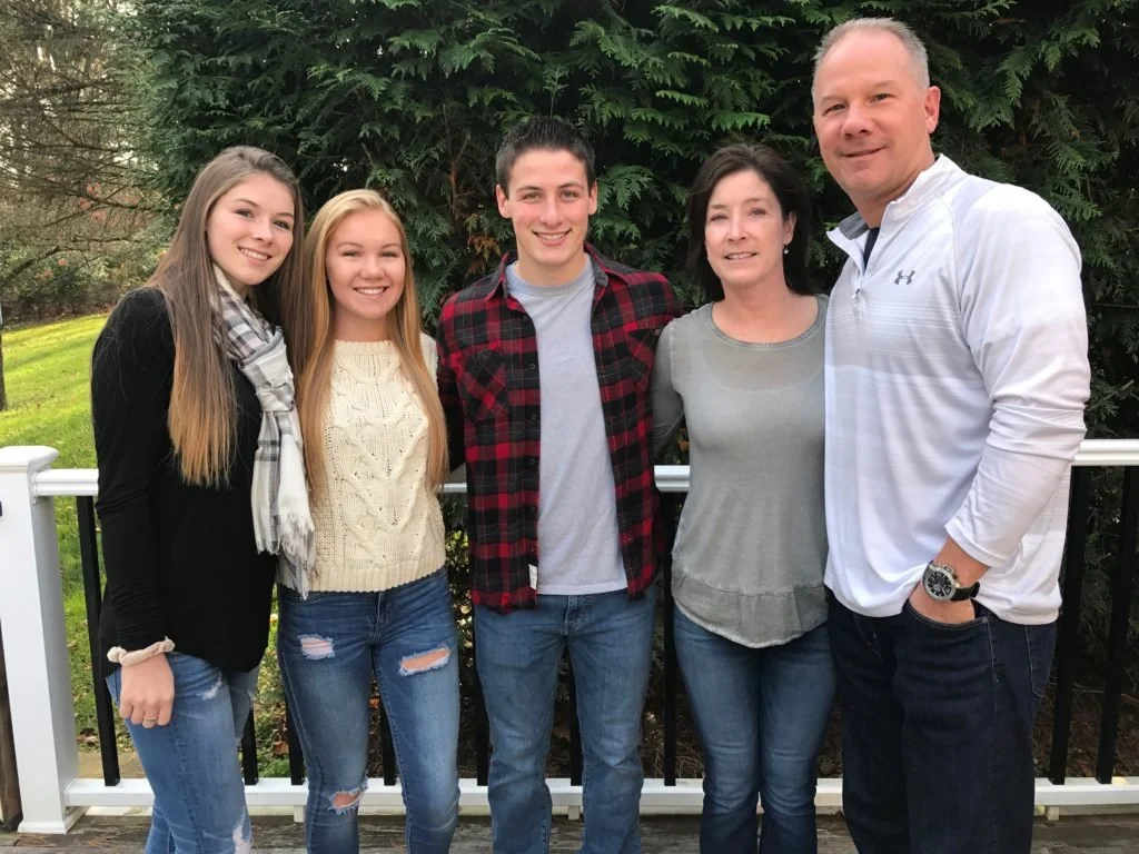 A family of six posing outdoors on a deck with greenery in the background, all smiling at the camera.