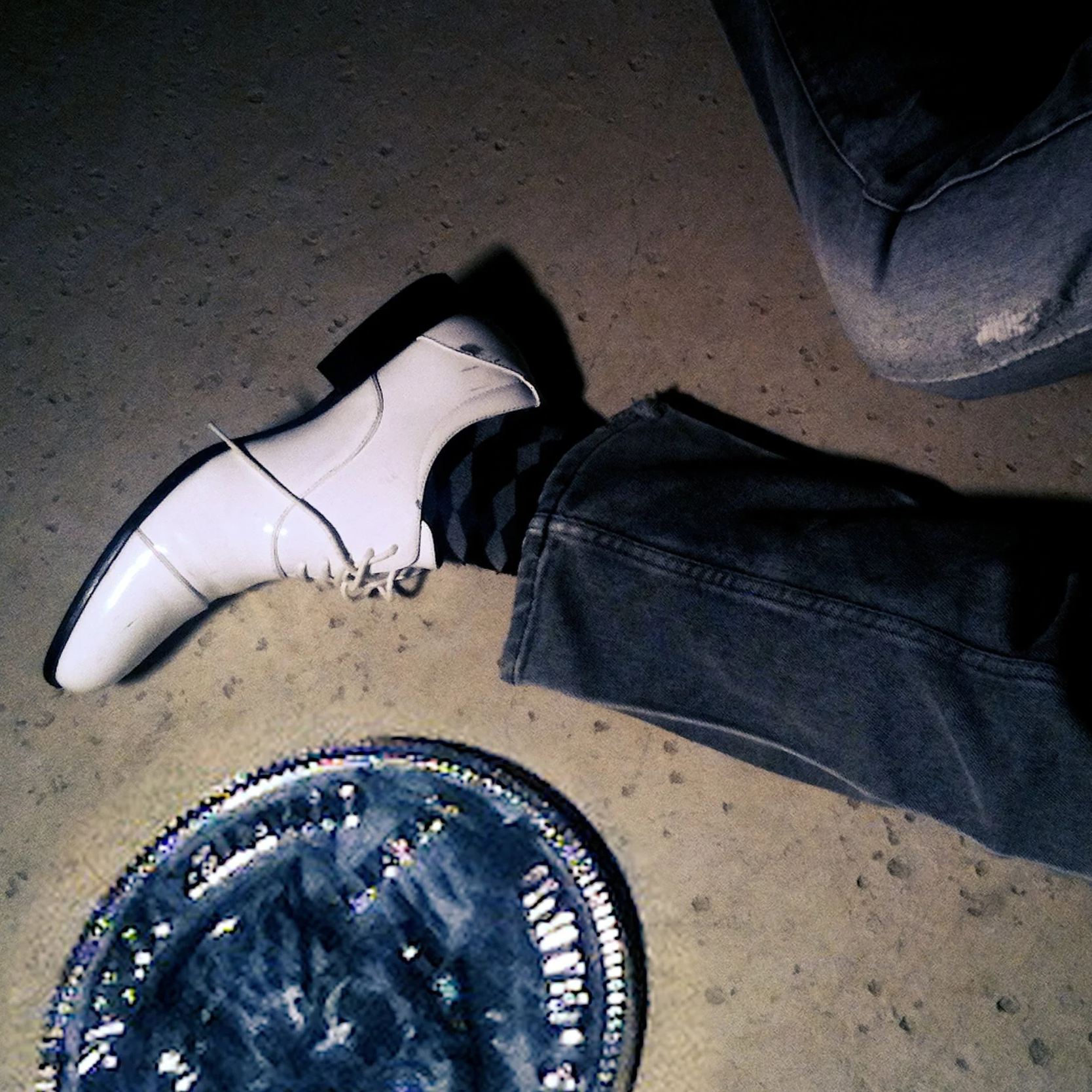 Close-up of a person's right foot wearing a shiny white dress shoe with black soles, paired with dark striped stockings and denim jeans. The background shows a textured floor and part of another person's leg in faded jeans.