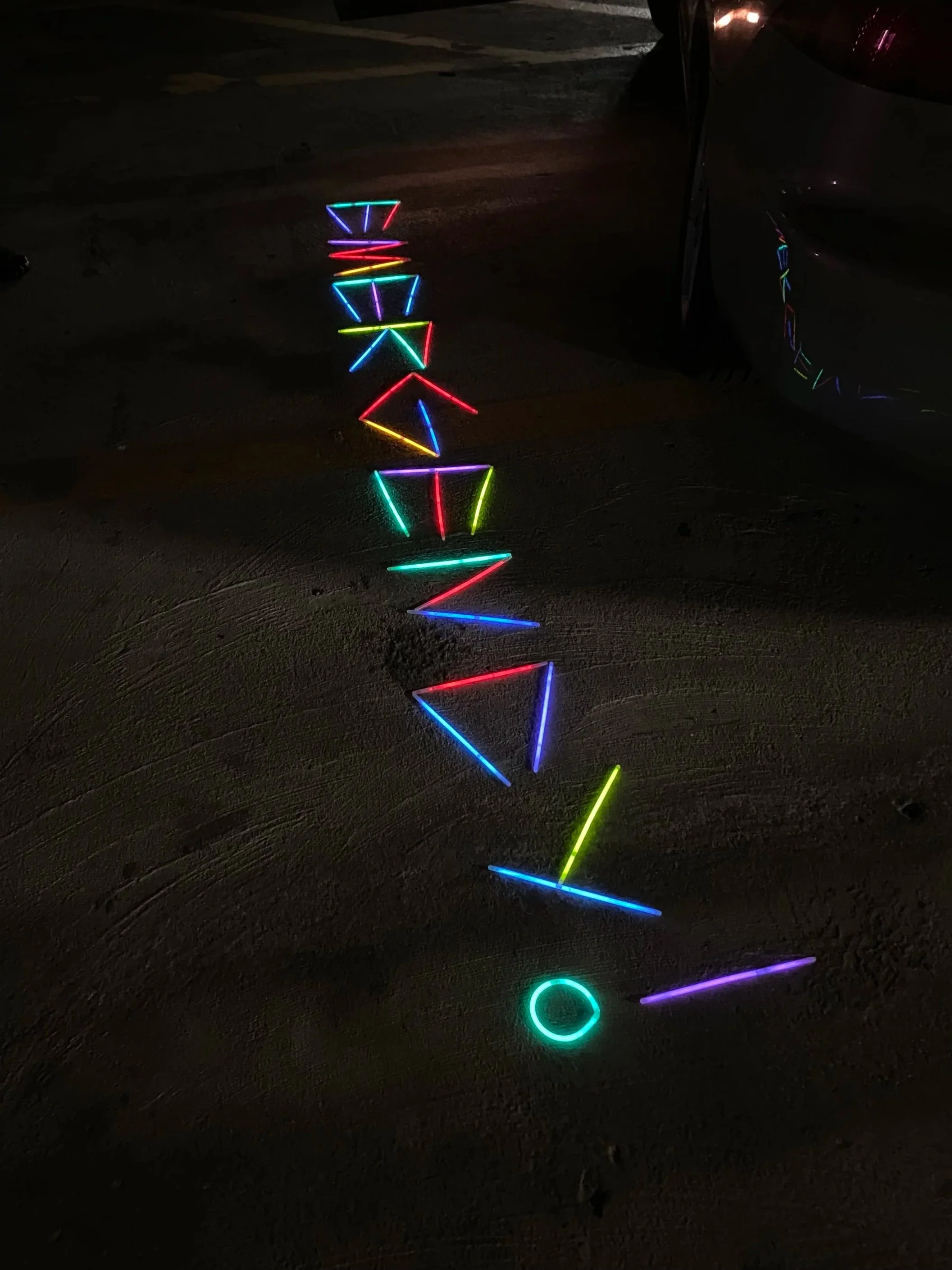 Colorful neon lights arranged to form letters spelling 'Emergency' on the ground in a parking lot at night, with a reflection of the lights on a parked car.