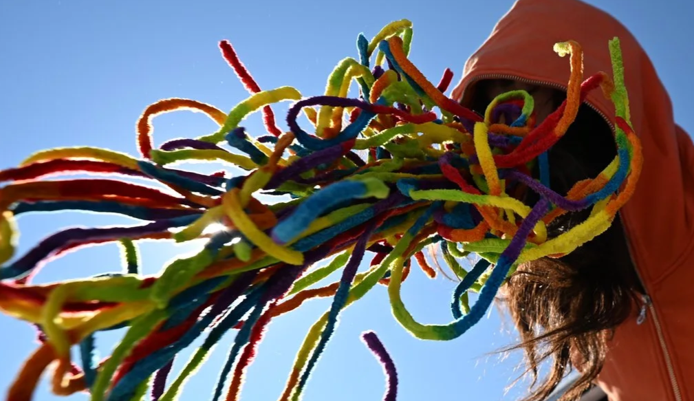 Person wearing a hoodie with colorful yarn strands streaming towards the camera against a blue sky.