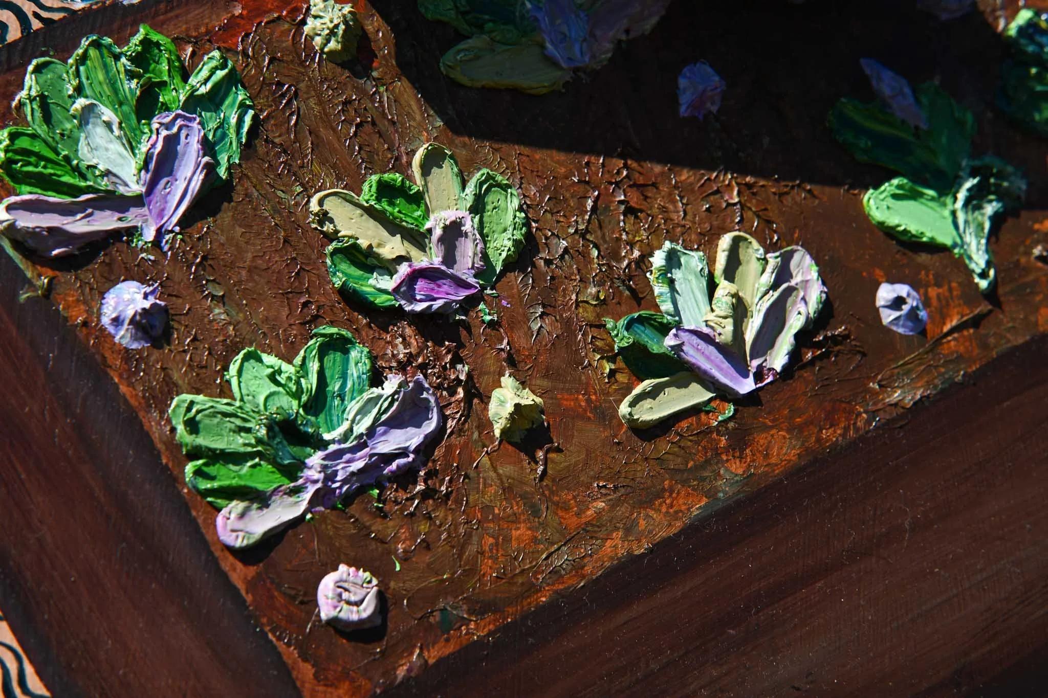 Close-up of textured, colorful, thickly applied oil paint on a wooden canvas, depicting abstract floral or leaf shapes in shades of green, purple, and white.