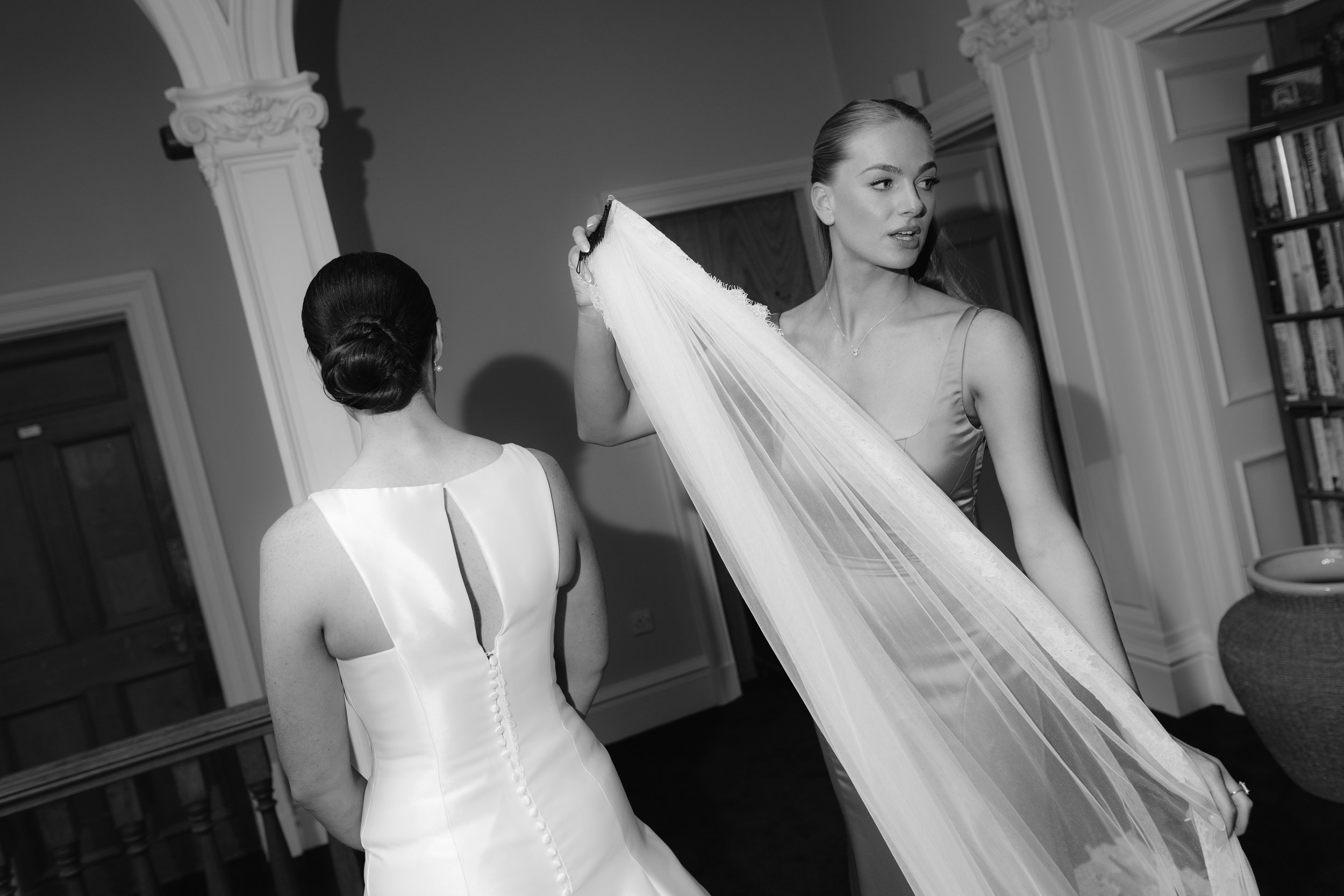 Two women, one in a wedding dress and the other holding a veil, inside a decorated room with bookshelves and ornate molding.