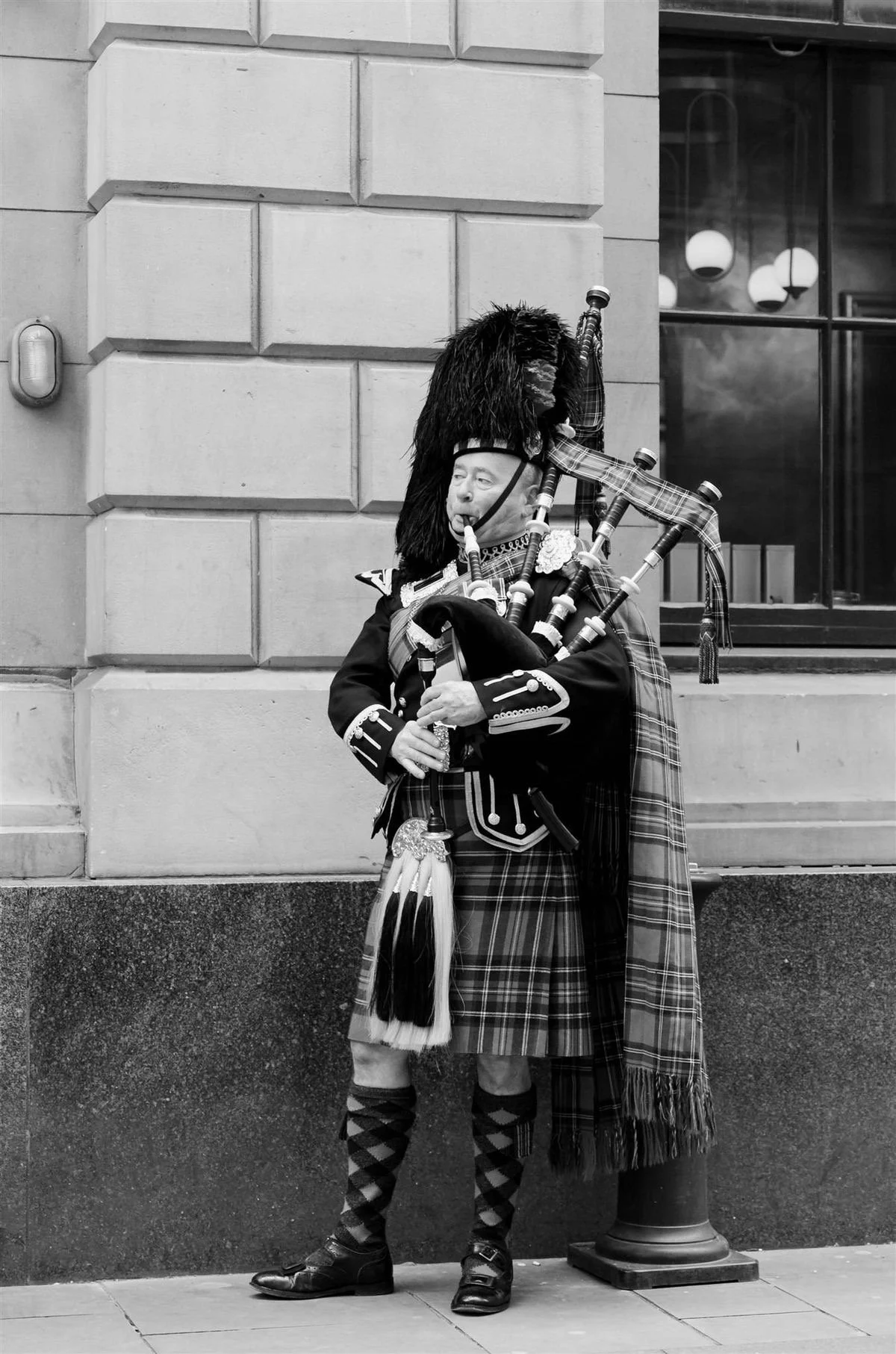 A man dressed in traditional Scottish attire playing bagpipes on a city sidewalk, standing next to a building with a window.