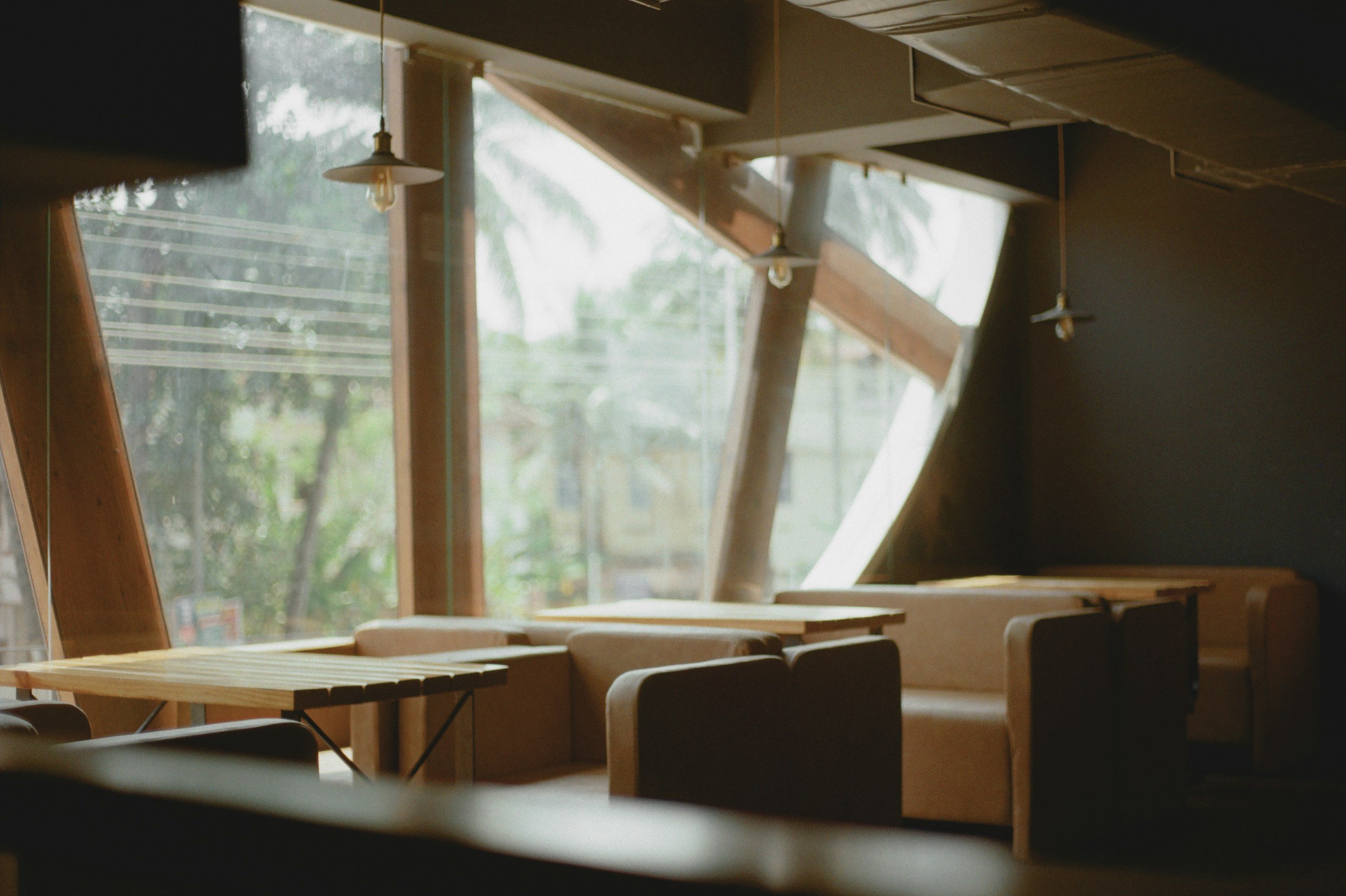 Interior of a cafe or restaurant with large windows, wooden furniture, and hanging light bulbs.
