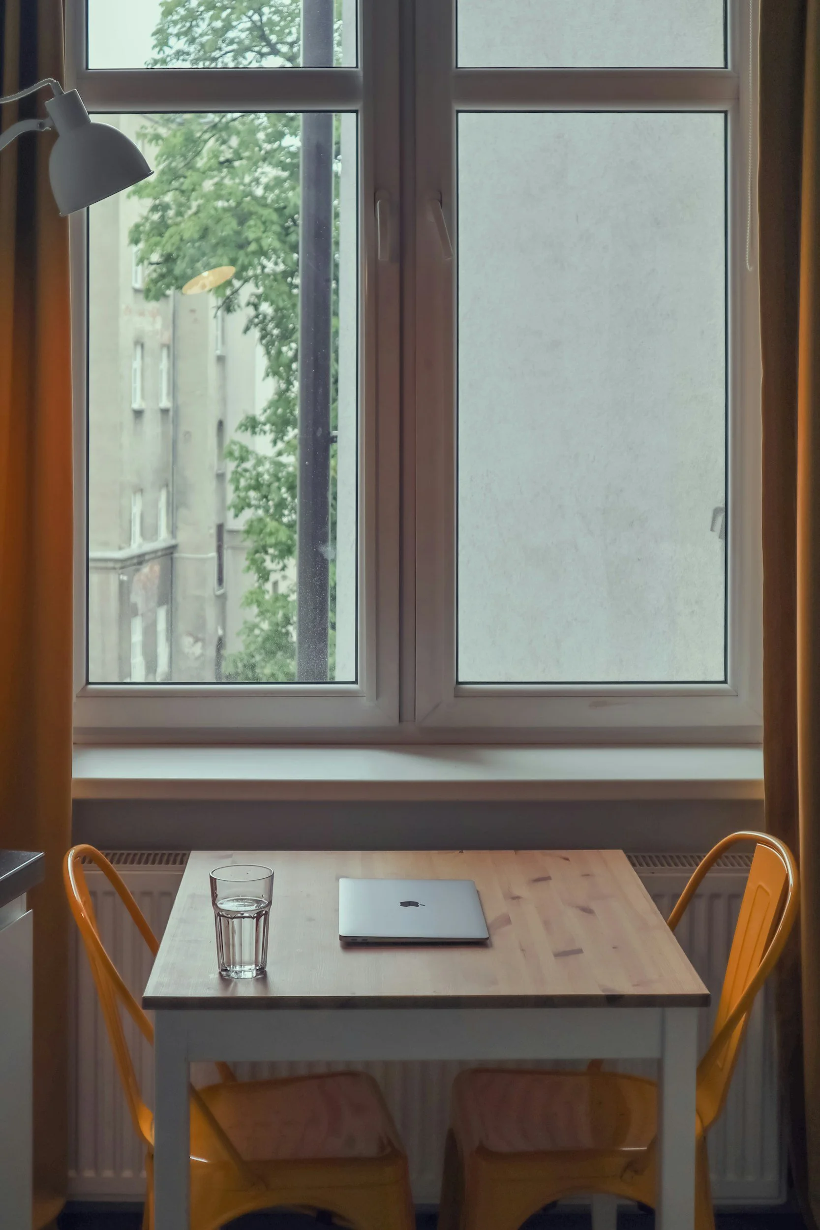 A wooden table with a closed silver laptop and a glass of water, flanked by two yellow chairs, set in front of a window with trees outside and orange curtains on the sides.