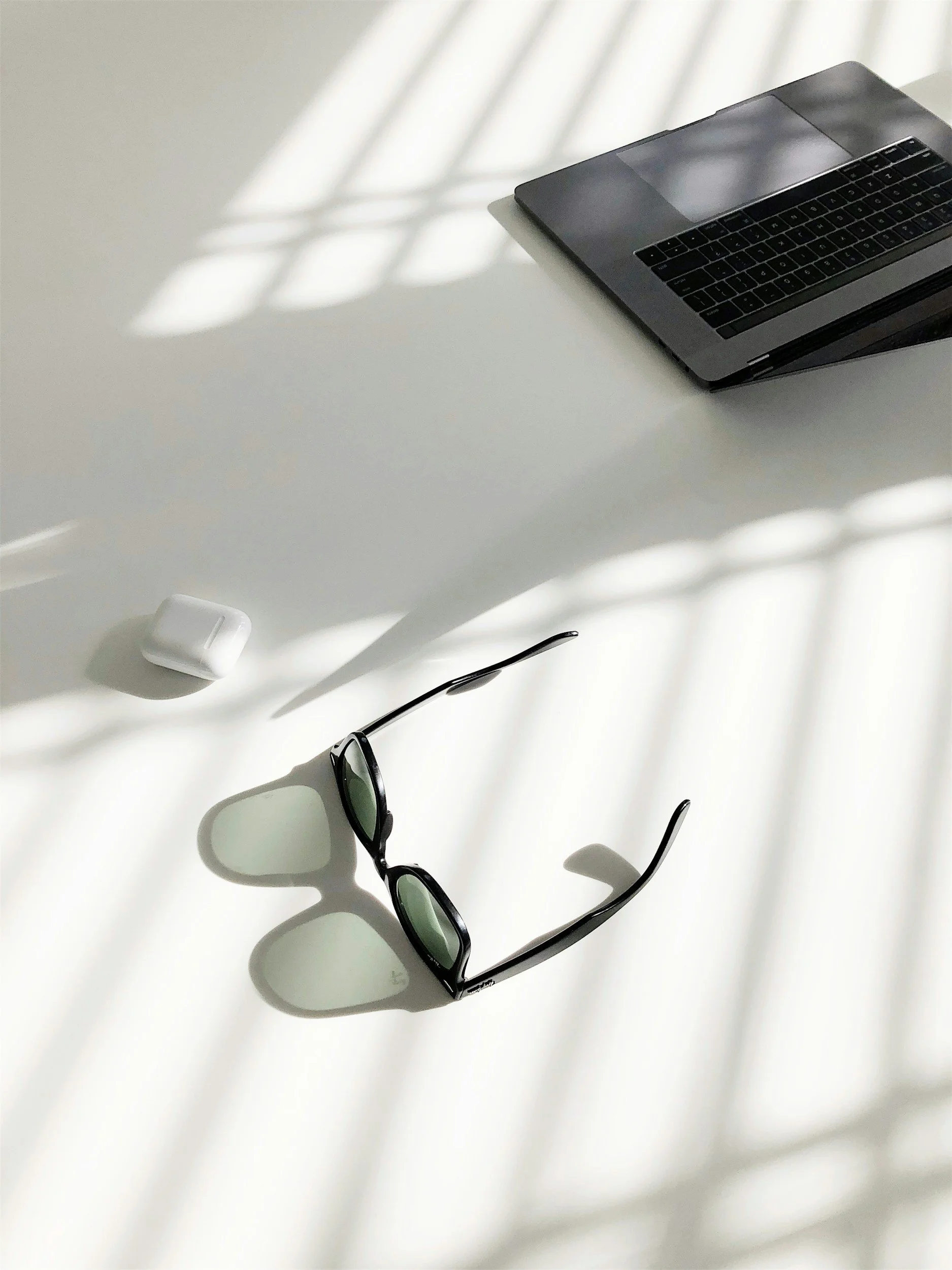 Sunlight casting shadows of a window grid on a white surface, with a black laptop, white wireless earbuds, and black sunglasses placed on the surface.