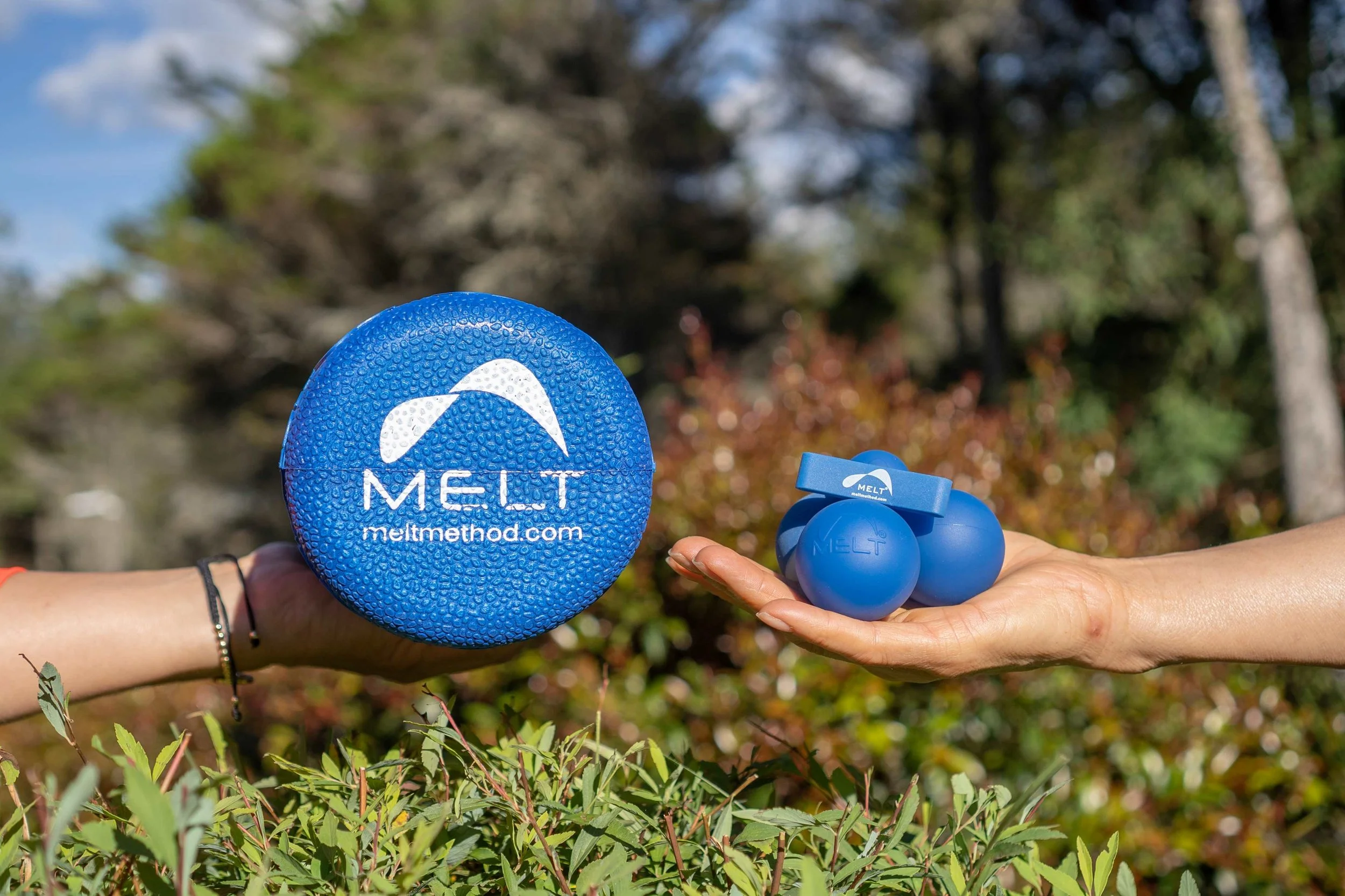 Close-up of two hands holding blue wellness products: one is a round pill box with the word 'MELT' and website 'meltmethod.com,' and the other is a small container with blue balls labeled 'MELT.' The background shows blurred autumn trees and a clear sky.