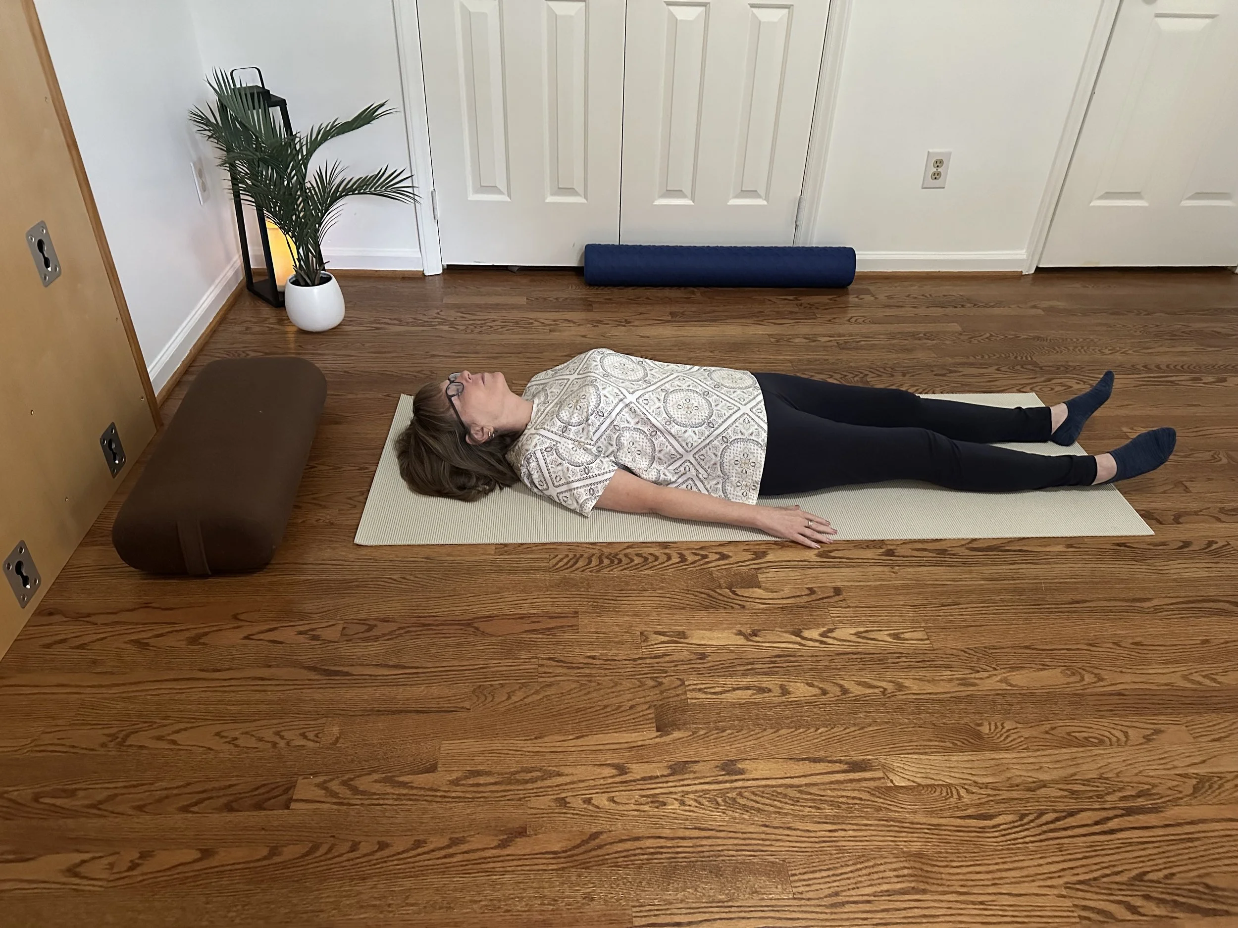 A woman lying on a yoga mat in a room, with a potted plant and a lamp nearby.