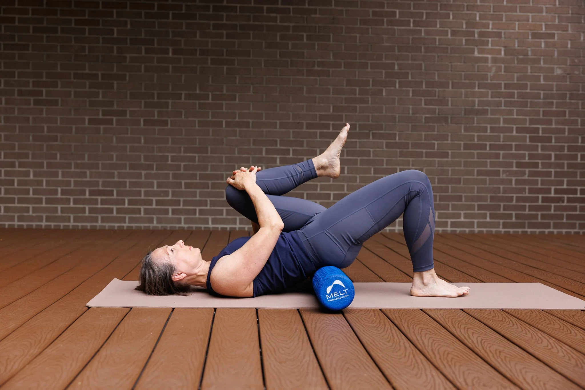 Senior woman practicing yoga on a mat with foam roller and massage ball, lying on her back and stretching her leg, indoors with a brick wall background.