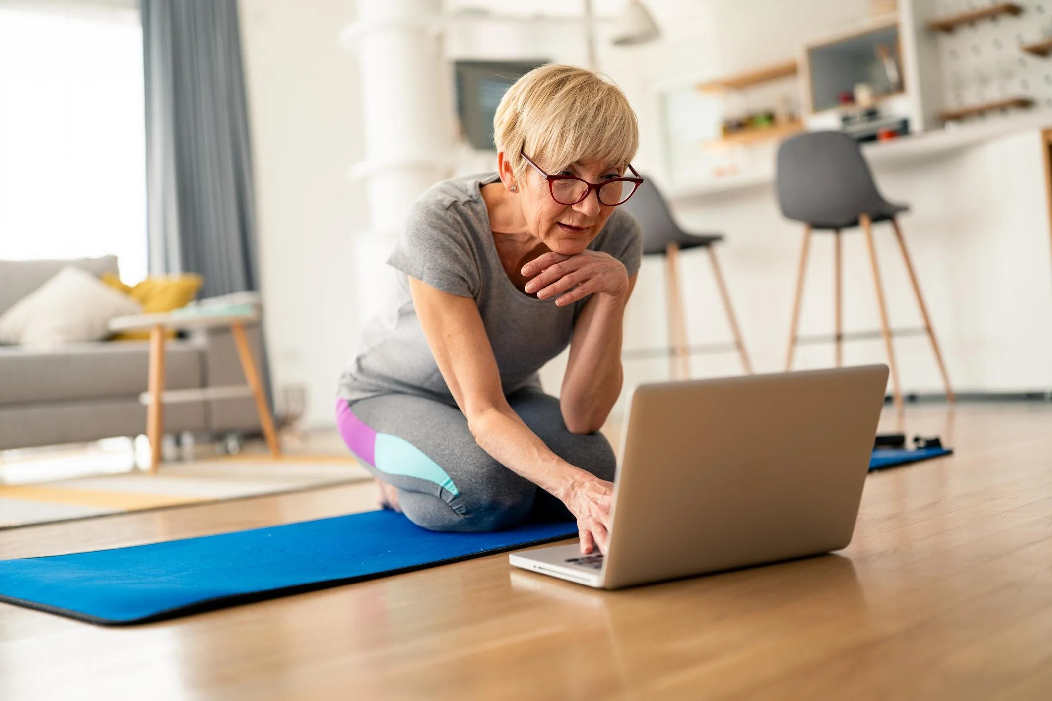 Older woman practicing yoga on a blue mat, kneeling and leaning forward while looking at a laptop, in a bright living room.