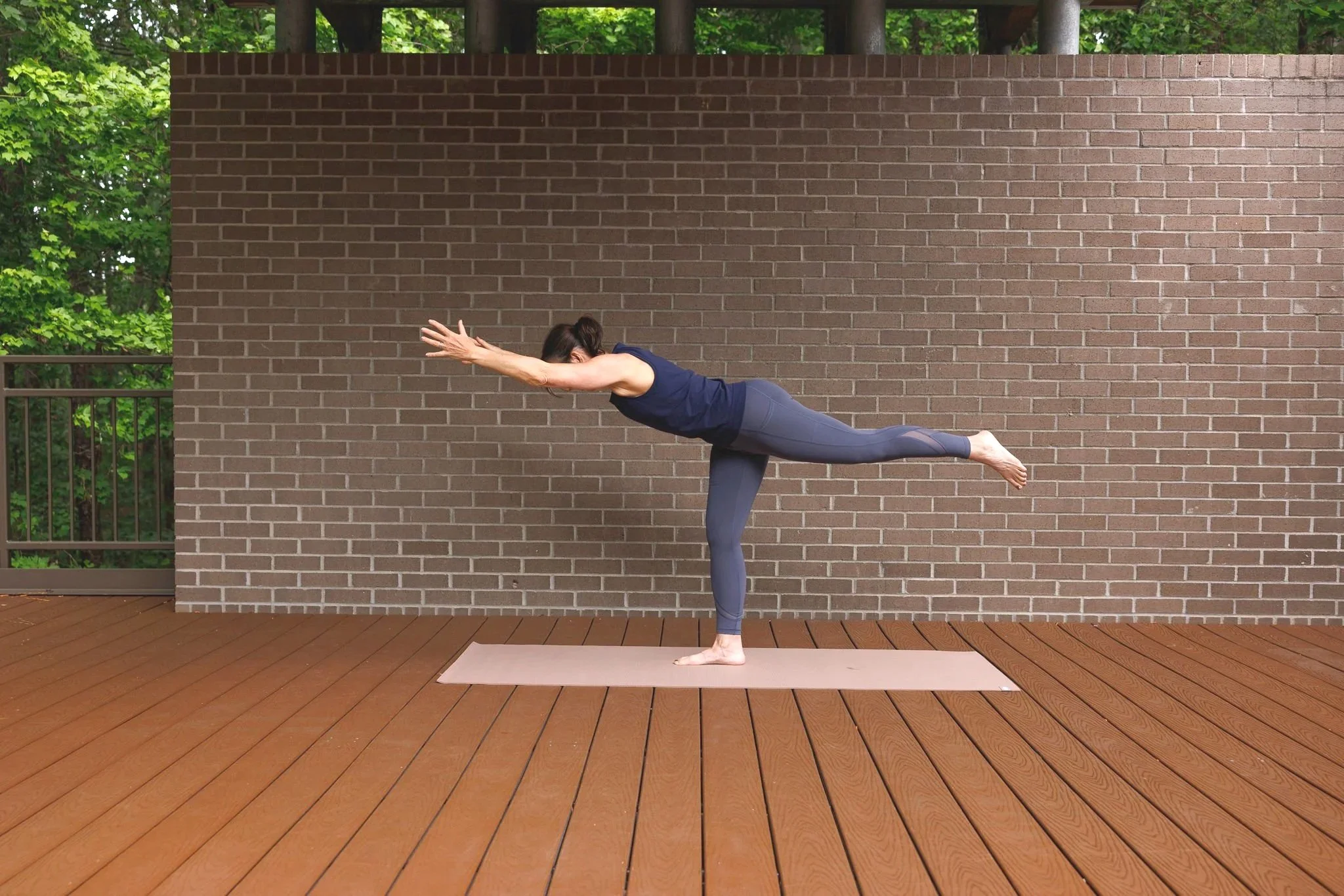 A woman practices yoga outdoors on a wooden deck. She is in a balancing pose, standing on one leg with the other leg extended behind her, and arms stretched forward. A brick wall and green trees are in the background.