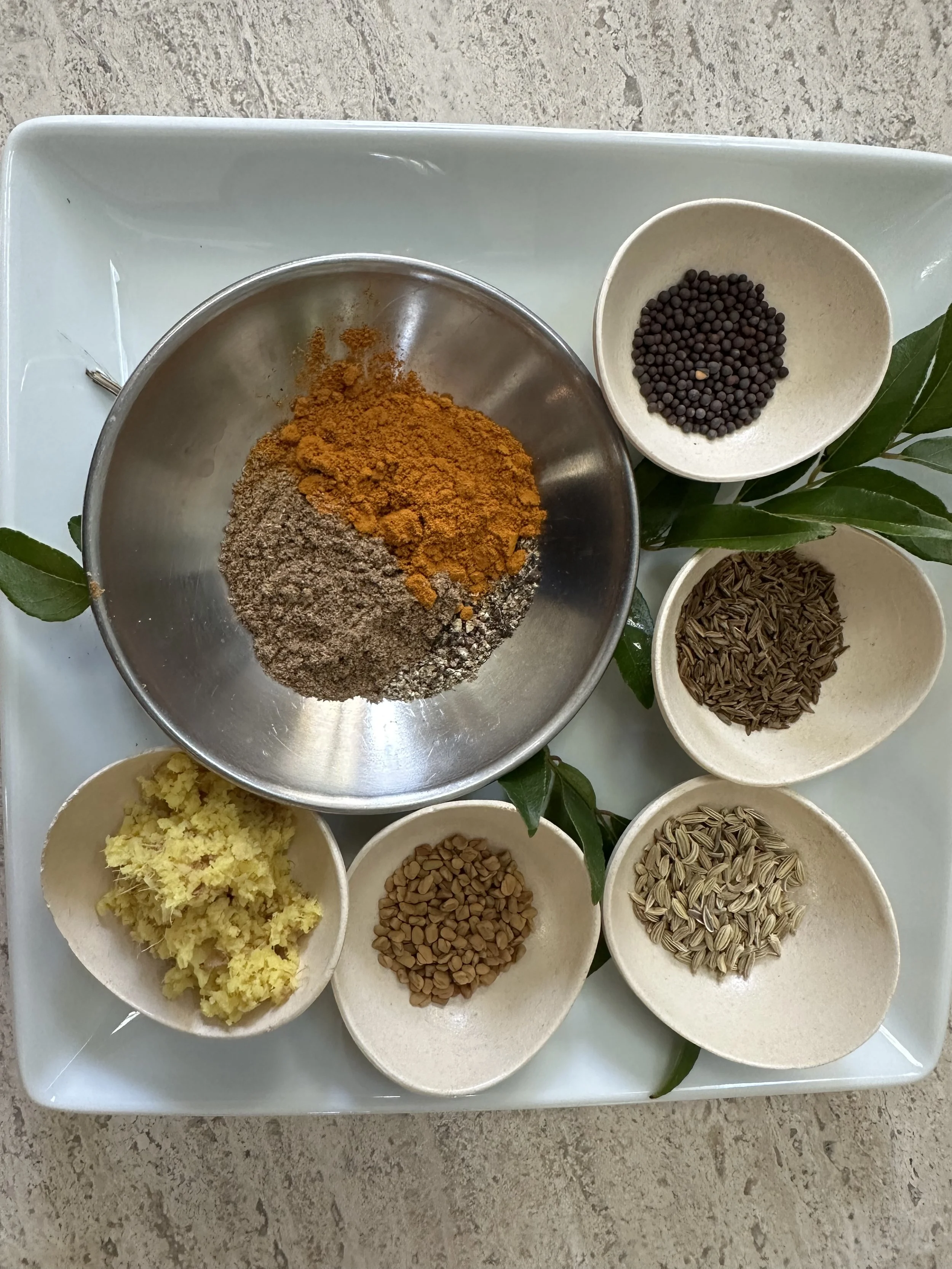 Assorted spices and seeds on a white tray, including yellow minced ginger, cumin seeds, black peppercorns, sunflower seeds, fennel seeds, and a spice blend in a metal bowl.