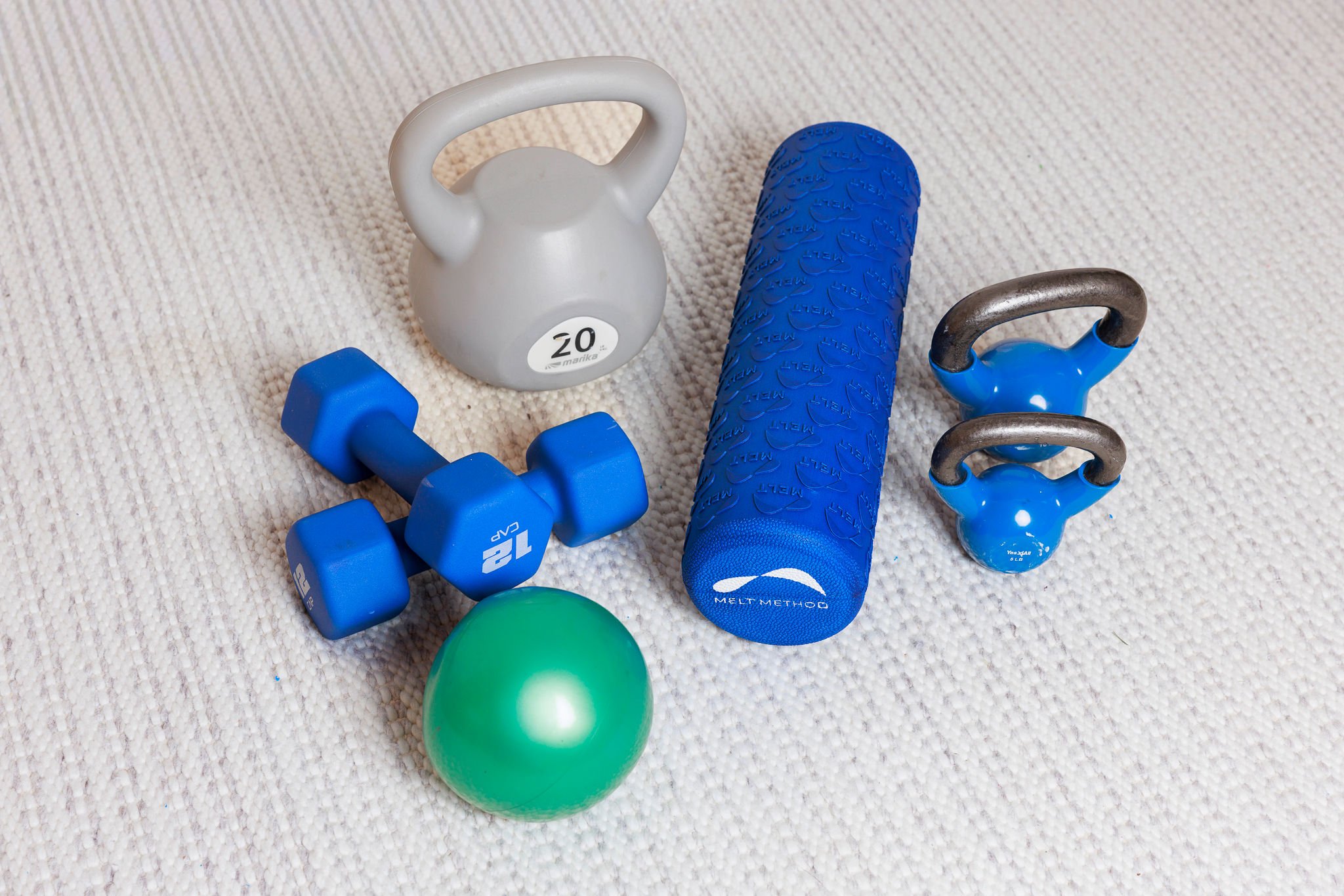 A collection of gym equipment on a beige carpet, including a white kettlebell, blue dumbbells, a blue foam roller, a small blue kettlebell, and a green exercise ball.