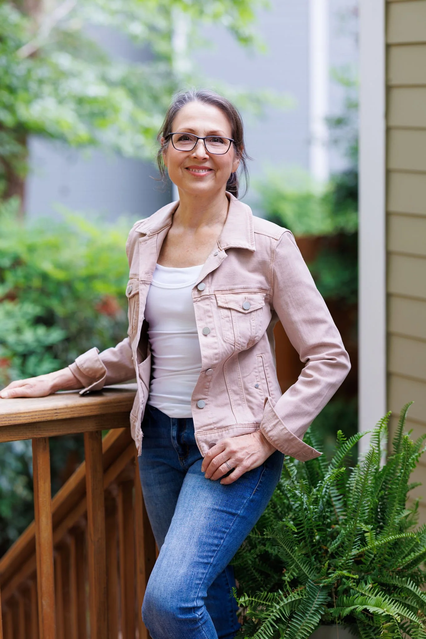 A woman with glasses standing outdoors on a wooden deck, leaning against a railing, surrounded by green plants and trees, smiling at the camera.