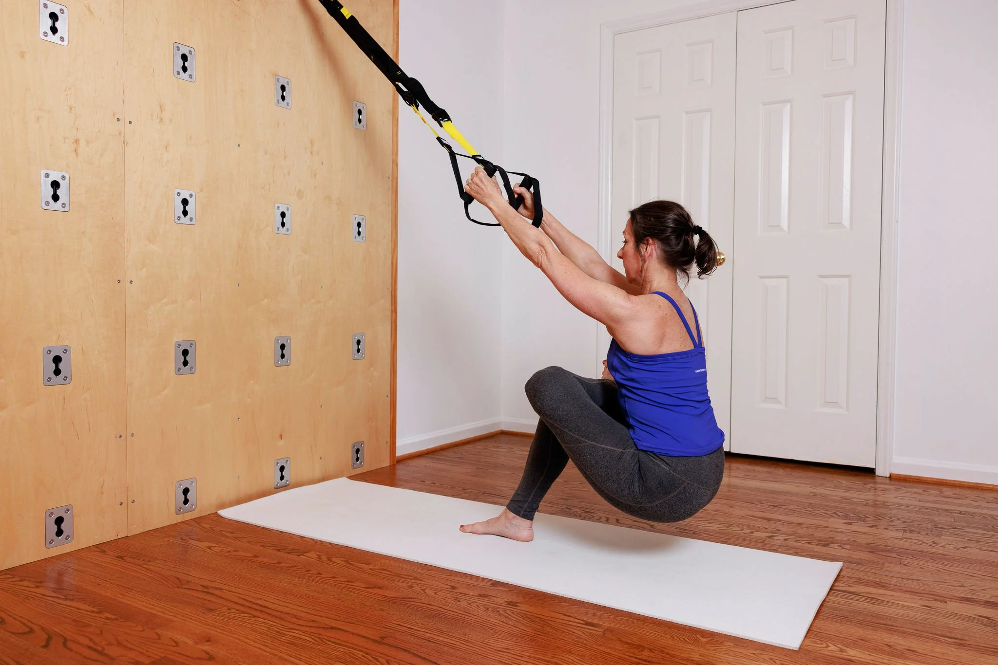 A woman exercises using suspension training equipment in a home workout space with a wooden wall and white closet doors.