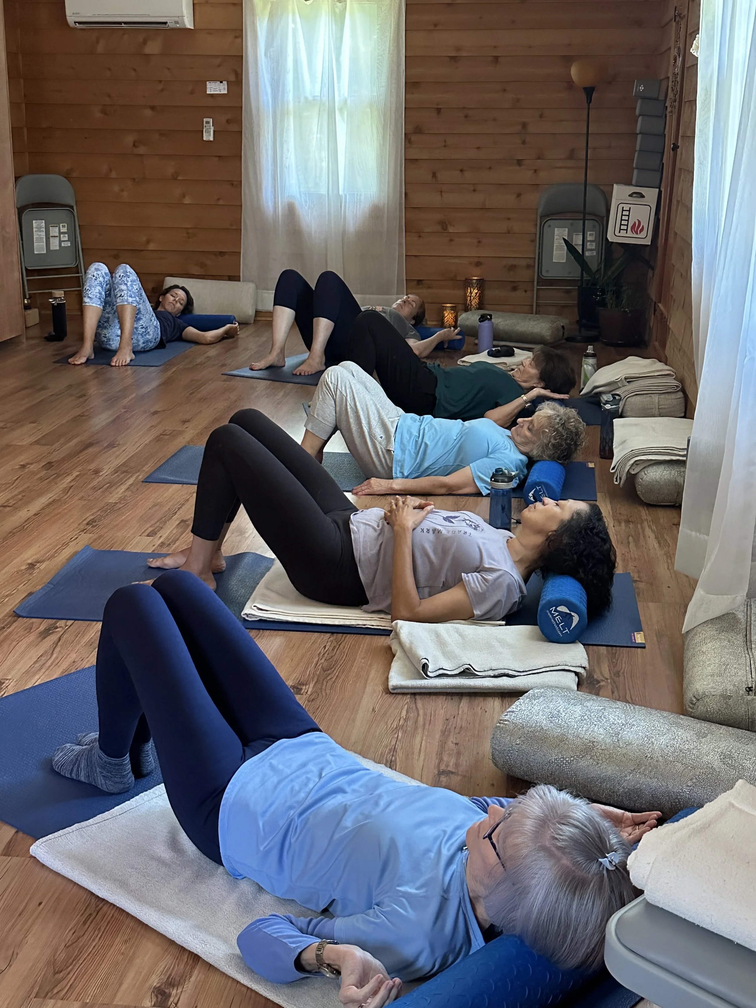 A group of people practicing yoga or meditation on mats and cushions in a cozy wooden room, lying on their sides or backs with eyes closed, surrounded by folded towels, water bottles, and lamps.